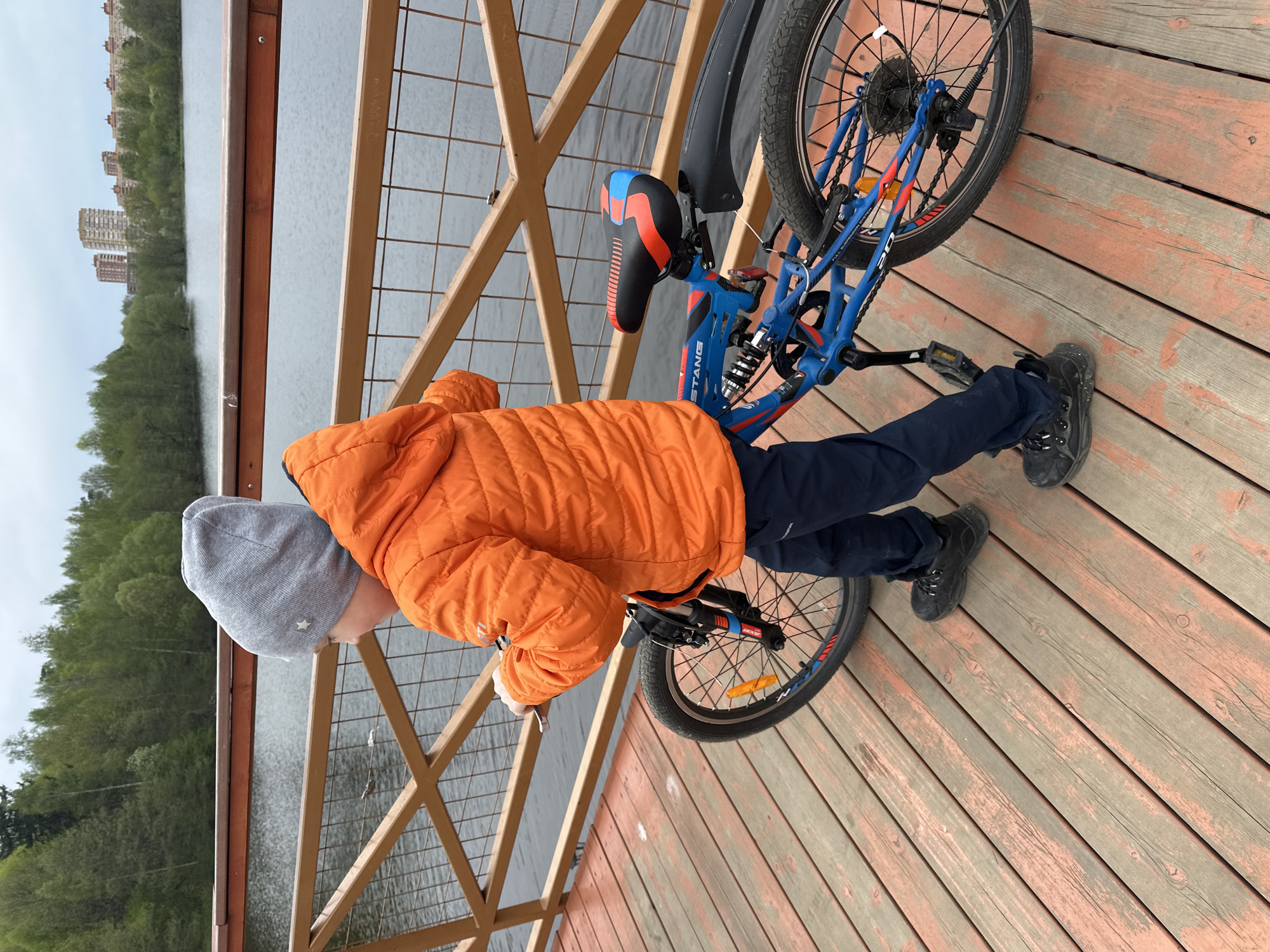 A boy in a gray hat and orange puffer jacket stands on a wooden deck, leaning over a railing, seemingly looking out at a body of water. A blue bicycle is propped against the railing next to him. The location appears to be a waterfront area in Grebnevo, Russia, based on the buildings and natural environment visible in the background. The wooden deck has a grid-like railing and is constructed of planks.