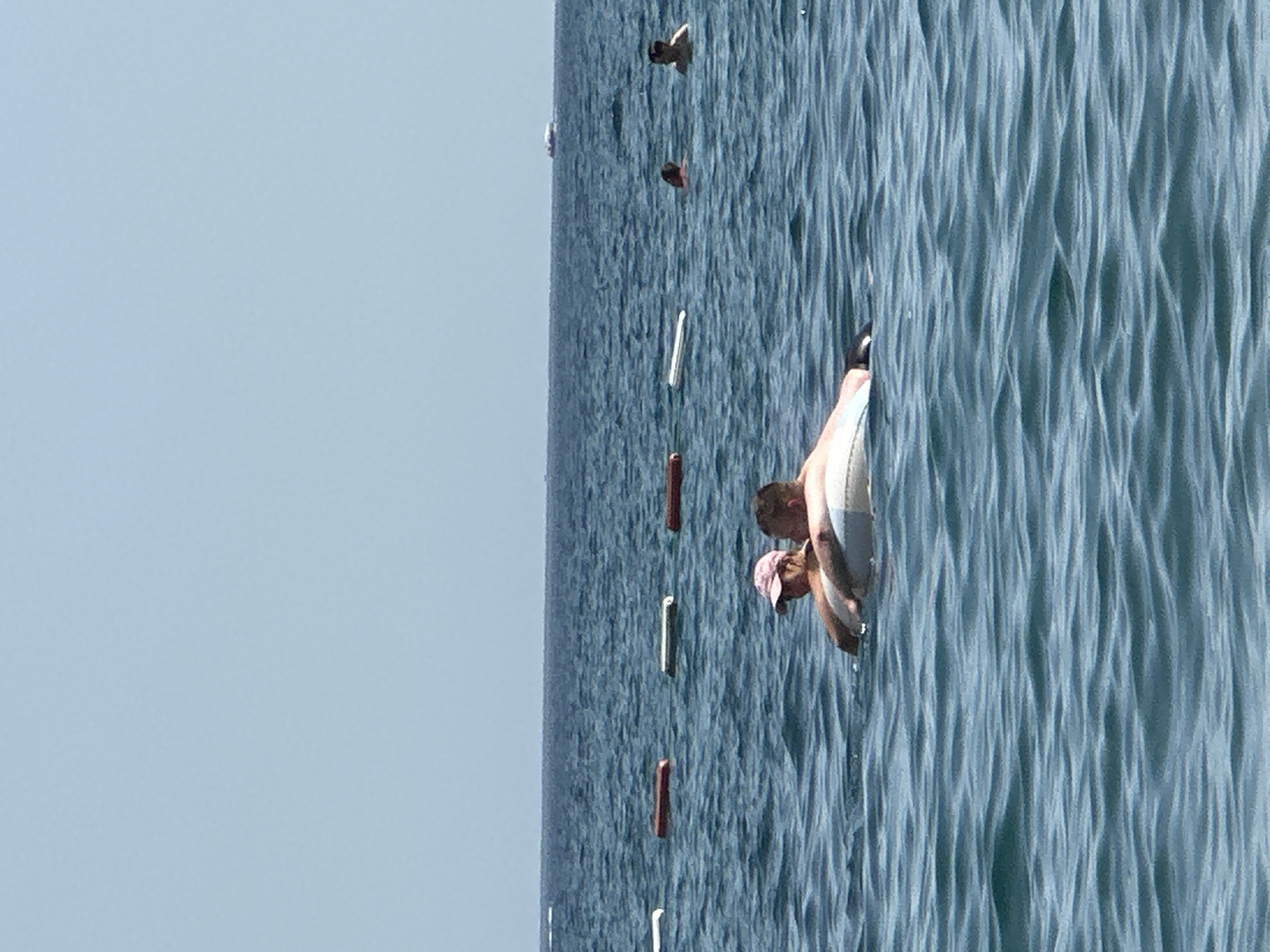 In the city of Boreti, Montenegro, two individuals are partially submerged in the blue, choppy waters of the Adriatic Sea. They are positioned centrally in the frame, embracing and resting on an inflatable ring. One individual, wearing a pink baseball cap and a dark top, is positioned in front, leaning into the other. The second individual, with short hair and a bare chest, is behind, with their arms around the front person. The water extends to the horizon, where it meets a pale blue sky. Several buoys, marked with alternating red and white or brown and white sections, are visible in a line across the middle ground of the image. In the distance, two other individuals are swimming, and a boat is visible on the far horizon. The lighting suggests it is daytime, likely a sunny day.