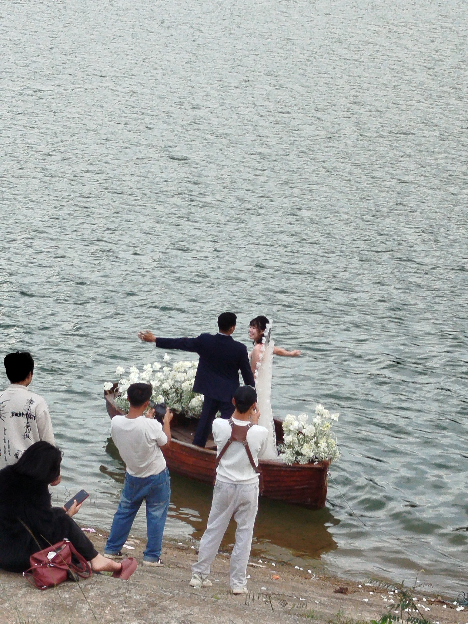 A wedding photoshoot is taking place on a body of water, likely Xuan Huong Lake in Da Lat, Vietnam, given the characteristic grey-green water and the style of clothing worn by the onlookers. The central subjects are a bride and groom in a small wooden boat adorned with white flowers. The groom, wearing a dark suit, is facing away from the viewer and towards the bride, with his right arm extended. The bride, in a white wedding dress, is facing towards the groom with her left arm extended.
Several other individuals are present, acting as photographers and spectators. On the shore, a person in a black outfit is seated, looking at a phone and holding a red bag. Another person standing near the shore is wearing a white t-shirt and blue jeans, holding a camera and seemingly taking a picture of the boat. Two other individuals in white t-shirts are also present near the boat, one appearing to take a photo and the other facing the couple. A person on the far left, wearing a white t-shirt with a graphic print, is standing and looking towards the boat. The water has a slightly choppy surface, and the shore is composed of dirt and scattered white petals. The overall atmosphere suggests a staged event with an audience.