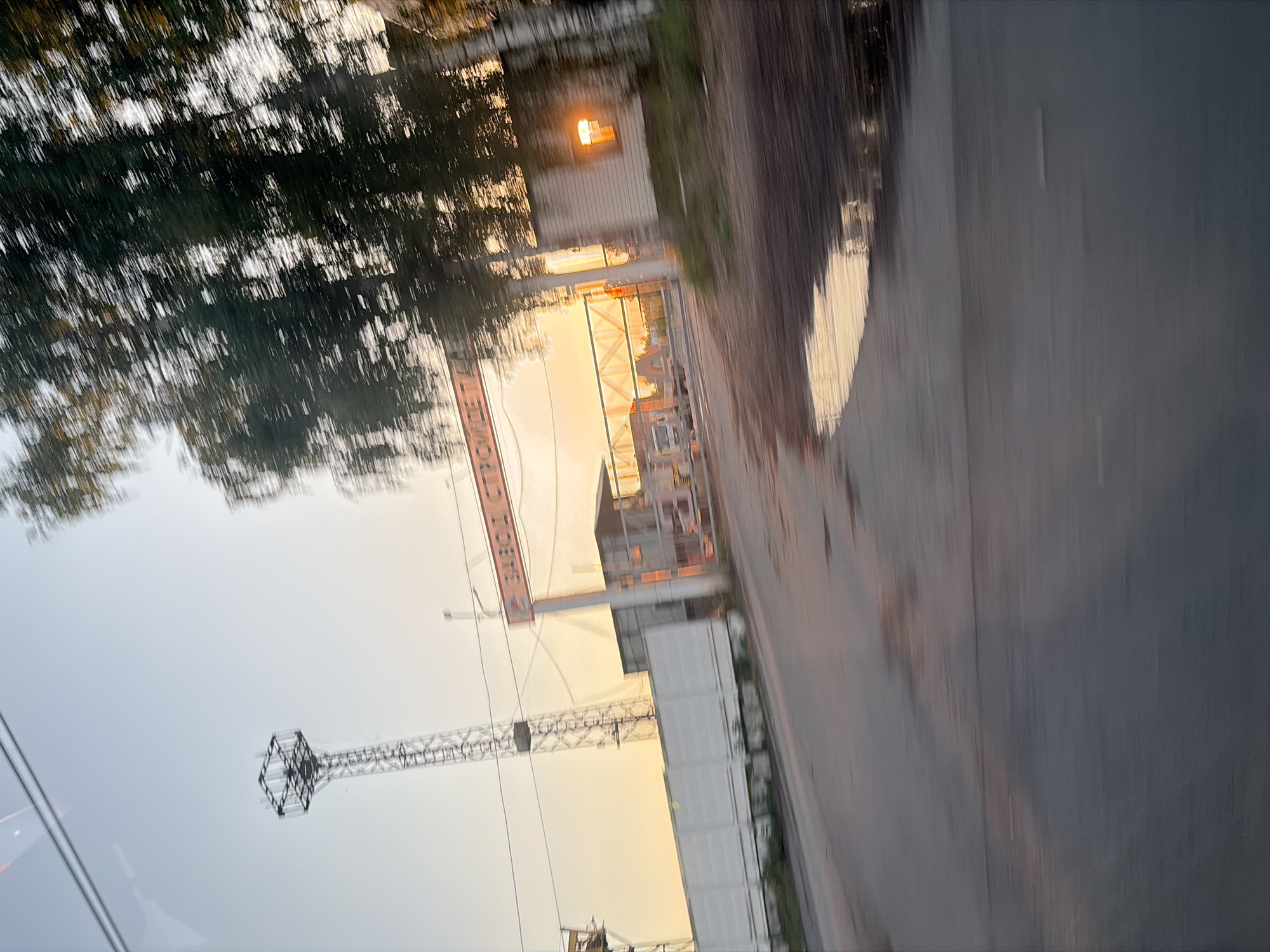 The image depicts a blurry, motion-distorted view of an industrial entrance at dusk or dawn. A tall, metal lattice tower stands to the left, and to the right, a gate with a sign that reads "ЗАВОД СТРОЙМАТЕРИАЛЫ" (meaning "Building Materials Plant"). Beyond the gate, there are buildings and structures, with a warm light emanating from a window on the right side. Trees with blurred foliage are visible in the foreground and mid-ground. The ground in front of the entrance is wet, with puddles reflecting the sky. The overall impression is of an industrial facility in a somewhat desolate or rural setting, captured during a moving vehicle's passage.