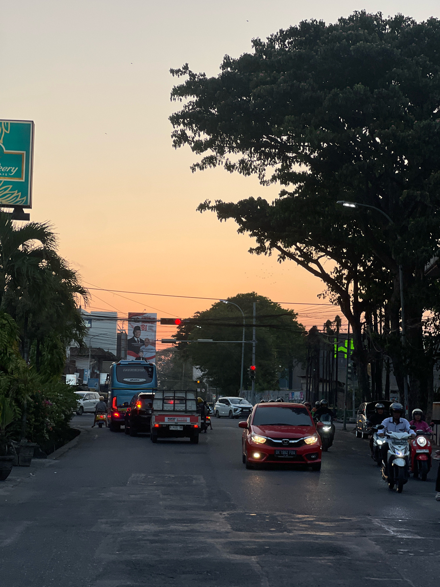 The image depicts a street scene in Kuta, Indonesia, during sunset. The sky is a gradient of warm orange and yellow hues. In the foreground, a paved road shows some wear and tear, with visible lane markings. Traffic is moderate, with several vehicles including a bright red car, a blue bus, a white pickup truck, and multiple motorbikes. Some motorbikes have riders wearing helmets. To the left, a billboard with green and teal accents advertises "Bakery Cafe." Further down the street, a large billboard features a portrait of a man in traditional attire, with text including "81." Lush green trees with dense foliage line the right side of the street, their branches silhouetted against the sky. A streetlamp with a modern design is visible among the trees. Several utility poles and wires are also present in the background. A few birds can be seen in the sky. The overall impression is a busy urban environment at dusk.
