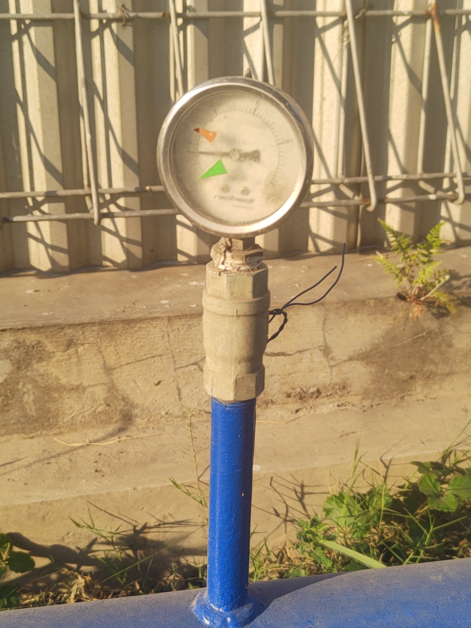 A pressure gauge on a blue pipe is positioned outdoors, against a backdrop of a corrugated metal fence and a concrete surface. The gauge face is circular with markings and a needle indicating a pressure reading. A thin black wire is attached to the pipe below the gauge. Green vegetation grows in patches around the base of the pipe and on the concrete. The lighting suggests it is daytime, casting shadows from the fence onto the concrete. The environment appears to be an industrial or utility area. The specific location in the city of Raci, Indonesia, is not discernible from the visual information provided in the image.