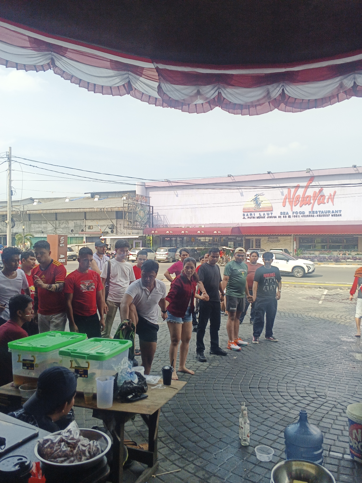 The image depicts an outdoor scene in front of a restaurant in the City of Medan, Indonesia, identified by the sign reading "Nelayan Sea Food Restaurant." A group of people, primarily young men and some women, are standing in a line, holding hands, and appear to be participating in a game or activity. They are dressed in casual attire, including t-shirts, shorts, and pants. Some individuals have their faces painted, suggesting a festive or competitive atmosphere.
In the foreground, there is a wooden table with several items on it, including large green plastic containers, clear plastic cups, a black disposable cup, and a bowl filled with food items, possibly snacks or appetizers. A person wearing a dark shirt and blue jacket is seated behind the table, facing away from the camera, and appears to be watching the activity.
The ground is paved with interlocking bricks arranged in a circular pattern. To the right of the table, a large blue water jug is visible, along with a small white bottle and a plastic container. In the background, the restaurant building features a prominent "Nelayan" sign in orange lettering and a graphic of a boat with a fisherman. Further in the background, several vehicles are parked along the street, and other buildings are visible, suggesting an urban setting. The sky is clear and bright.
