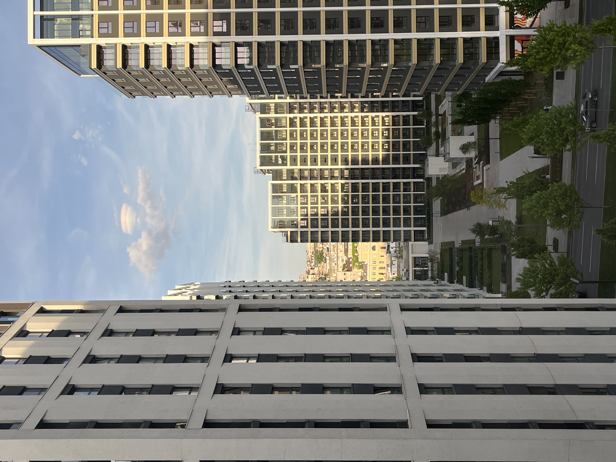 The image shows a view of buildings and the sky, likely taken from a high vantage point within a city, identifiable as Belgrade, Serbia, based on architectural styles present in the background.
Foreground: The lower part of a tall building with a gray facade and rectangular windows is prominent.
Middle Ground: There is a view of additional buildings, some with a grid-like window pattern, and a park-like area with trees and pathways.
Background: A cityscape is visible in the distance.
Sky: The sky is blue with some clouds.
The viewpoint is looking down between buildings, with a focus on the architecture and layout of the city.