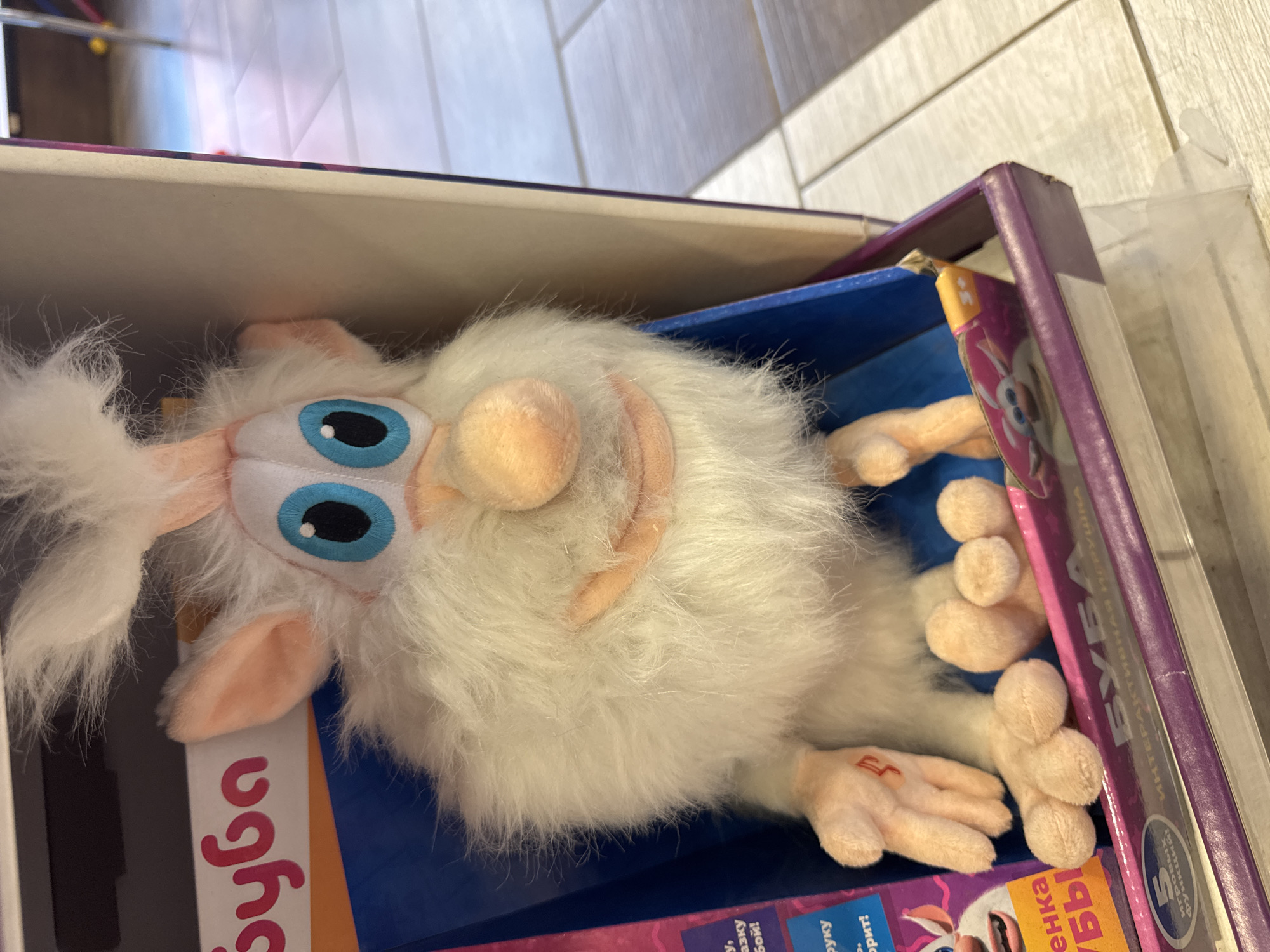 This image shows a close-up of a fluffy white toy with large blue eyes and an orange nose, sitting inside its packaging. The packaging is a cardboard box with purple and pink accents, and text in Russian is visible on it. The toy appears to be a character from a children's show or game. The background is blurred, but it appears to be an indoor setting with a tiled floor. The overall impression is that this is a product photo for a toy. There is no information to suggest this is in Marchugi, Russia.