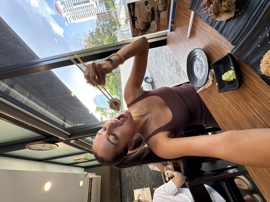 The image displays an interior view of a Japanese restaurant, looking outwards towards an urban environment.
A young adult female with dark hair tied in a ponytail is centrally positioned, oriented towards the camera. She is wearing a brown tank top and a silver necklace with an "S" pendant. Her mouth is open. Her left hand, adorned with a watch and multiple beaded bracelets, holds a piece of sushi with wooden chopsticks, extended towards her open mouth. Her right forearm is visible in the lower left foreground, suggesting a first-person perspective or selfie.
The female subject is seated at a wooden table with a distinct grain pattern. On the table, in the foreground, are several items: a small, empty, dark-colored round plate; a small, square, dark-colored dish containing a portion of green wasabi; and two larger, rectangular, dark-colored plates holding sushi rolls. One sushi roll visible on a rectangular plate is topped with sesame seeds. A white paper wrapper for chopsticks is also present on the table. A menu board displaying food items and Thai text is visible on the table near the window.
In the background, to the left, another individual, a male wearing a white t-shirt and a baseball cap, is partially visible seated at another table. The wall behind him is dark and textured. An air conditioning unit and a circular wall clock are mounted on this wall.
Through a large window, the outdoor urban environment is visible. A blue circular sign with white text, clearly legible as "SUSHI HANA," is prominent. A white four-door sedan is parked on a paved surface outside. Green foliage from trees is visible. In the distant background, several modern high-rise buildings are visible against a blue sky with scattered clouds.
Based on the visible "SUSHI HANA" restaurant branding, the presence of Thai script on the menu, and the characteristic urban architecture, the location is identified as a Sushi Hana restaurant within the city of Bangkok, Thailand.