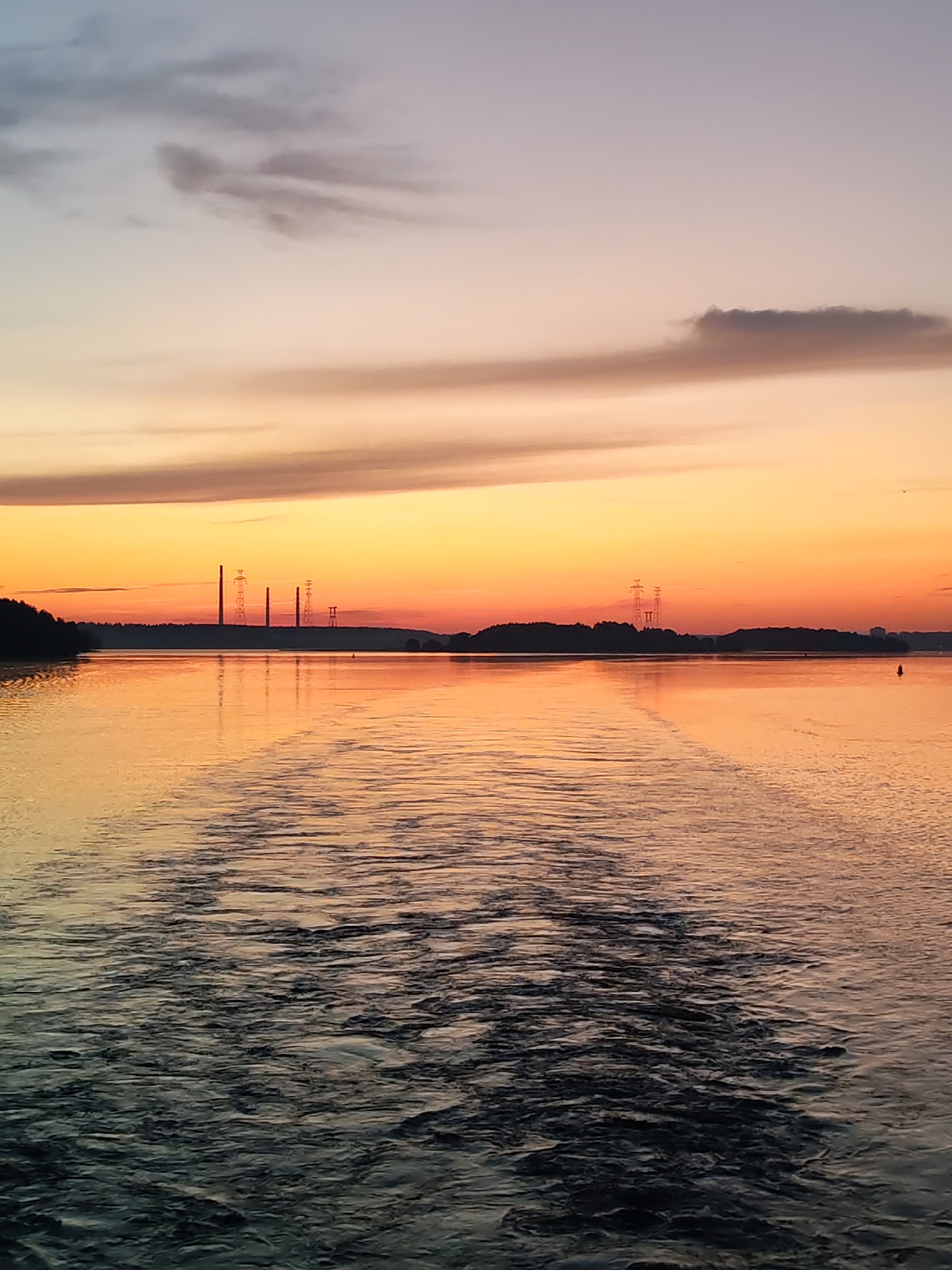 A vertical, medium shot captures a body of water during a sunset. The sky transitions from a pale blue at the top to vibrant oranges and yellows near the horizon, with scattered, wispy clouds. Silhouetted against the colorful sky are trees lining the distant shore, punctuated by several tall, industrial smokestacks and power lines. The water in the foreground is textured with ripples and waves, reflecting the colors of the sky. A wake from a boat is visible, extending from the bottom center of the frame towards the horizon. The overall scene conveys a serene, atmospheric mood.