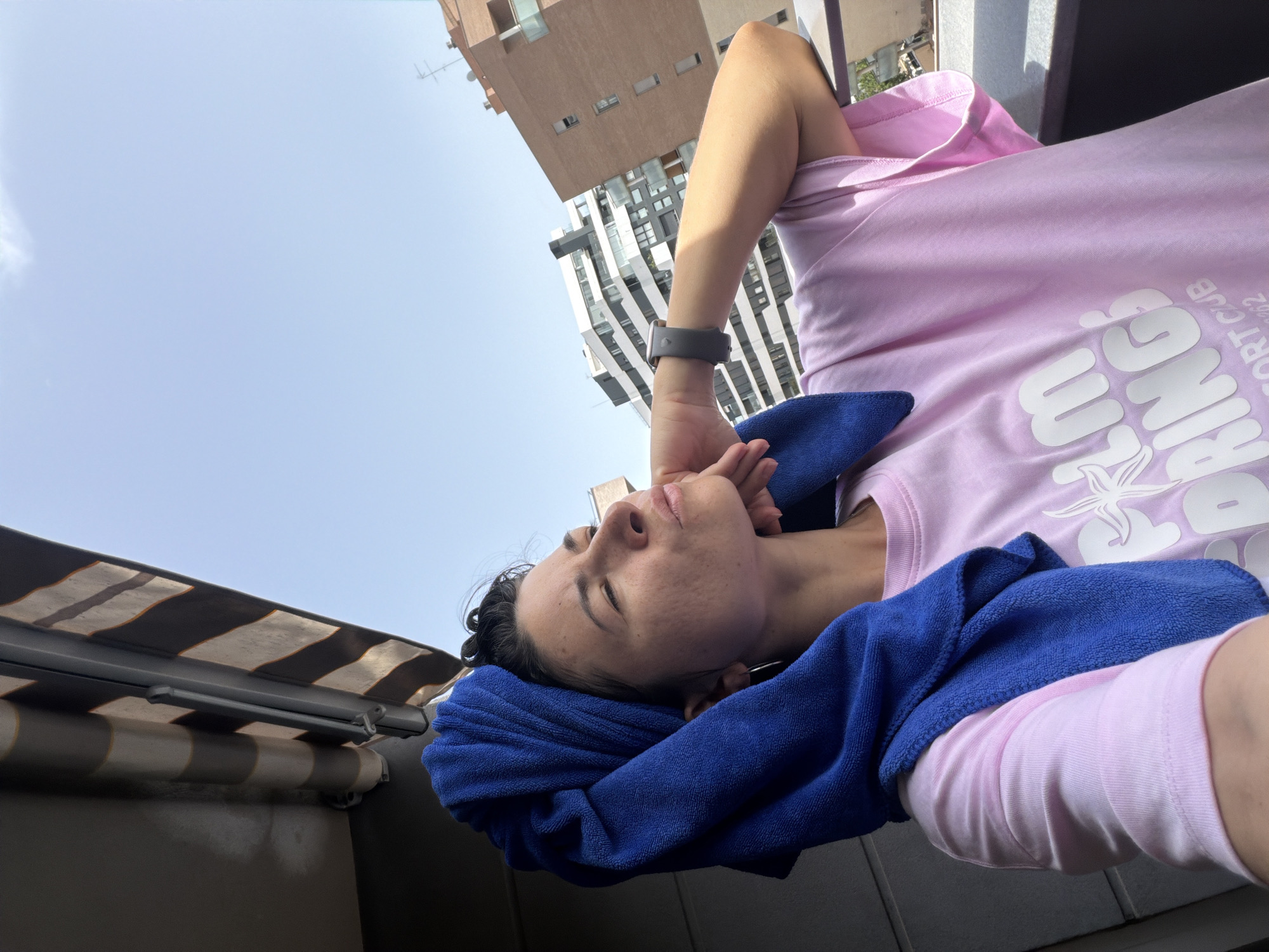 A woman stands on a balcony, facing right, with a blue towel wrapped around her head. She is wearing a light pink t-shirt with white text that reads "PALM SPRINGS RESORT CLUB 262". Her right arm is bent and resting on the railing, and her left hand is touching her chin. The background shows a clear blue sky with buildings and an awning. The lighting suggests it is daytime.