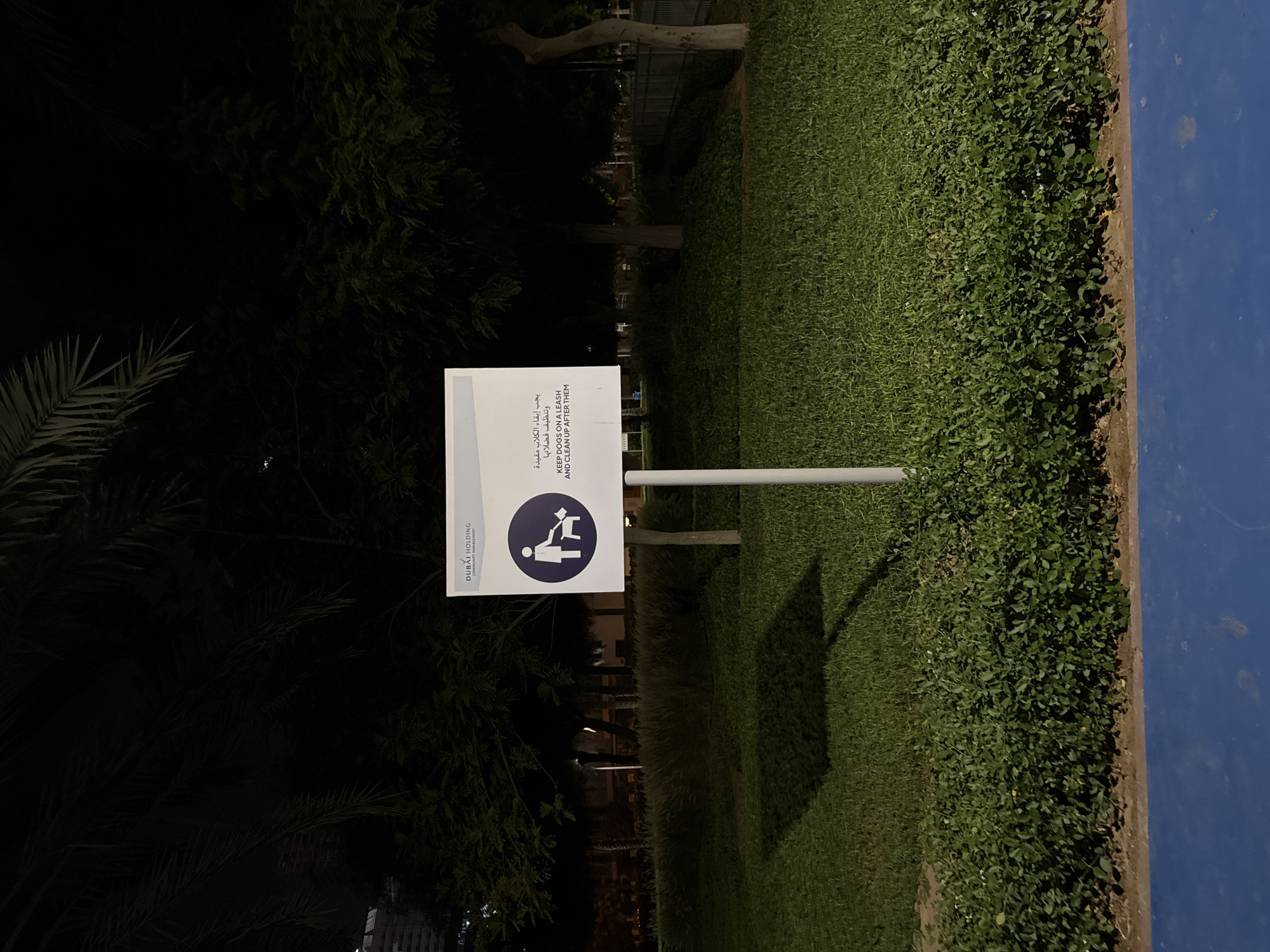 This media file shows a sign in a park at night. The sign is white with blue text and graphics. The text on the sign is in both Arabic and English and states: "DUBAI HOLDING COMMUNITY MANAGEMENT," "يجب إبقاء الكلاب مقيدة وتنظيف فضلاتها," and "KEEP DOGS ON A LEASH AND CLEAN UP AFTER THEM." There is also a blue circular graphic depicting a person picking up after a dog. The sign is mounted on a white pole. The setting is a grassy park with trees and a blue surface, possibly a track or sports area, visible on the right side of the frame. The image is dark, indicating it was taken at night. The lighting suggests artificial illumination within the park. The overall scene is peaceful and orderly, with the sign clearly communicating park regulations.