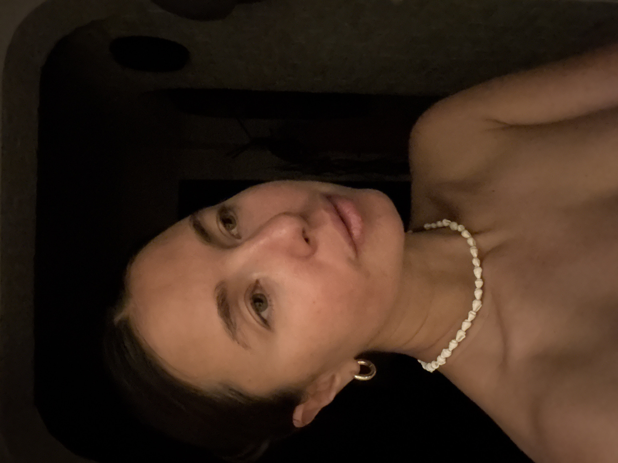 A close-up, dimly lit image depicts a woman's face and upper chest. She has light skin, brown hair pulled back, and is wearing a gold hoop earring in her left ear and a white beaded necklace. Her expression is neutral. The background is dark and appears to be a textured surface, possibly a bathtub or spa setting. There is no identifiable evidence to suggest the location is Kazan, Russia.