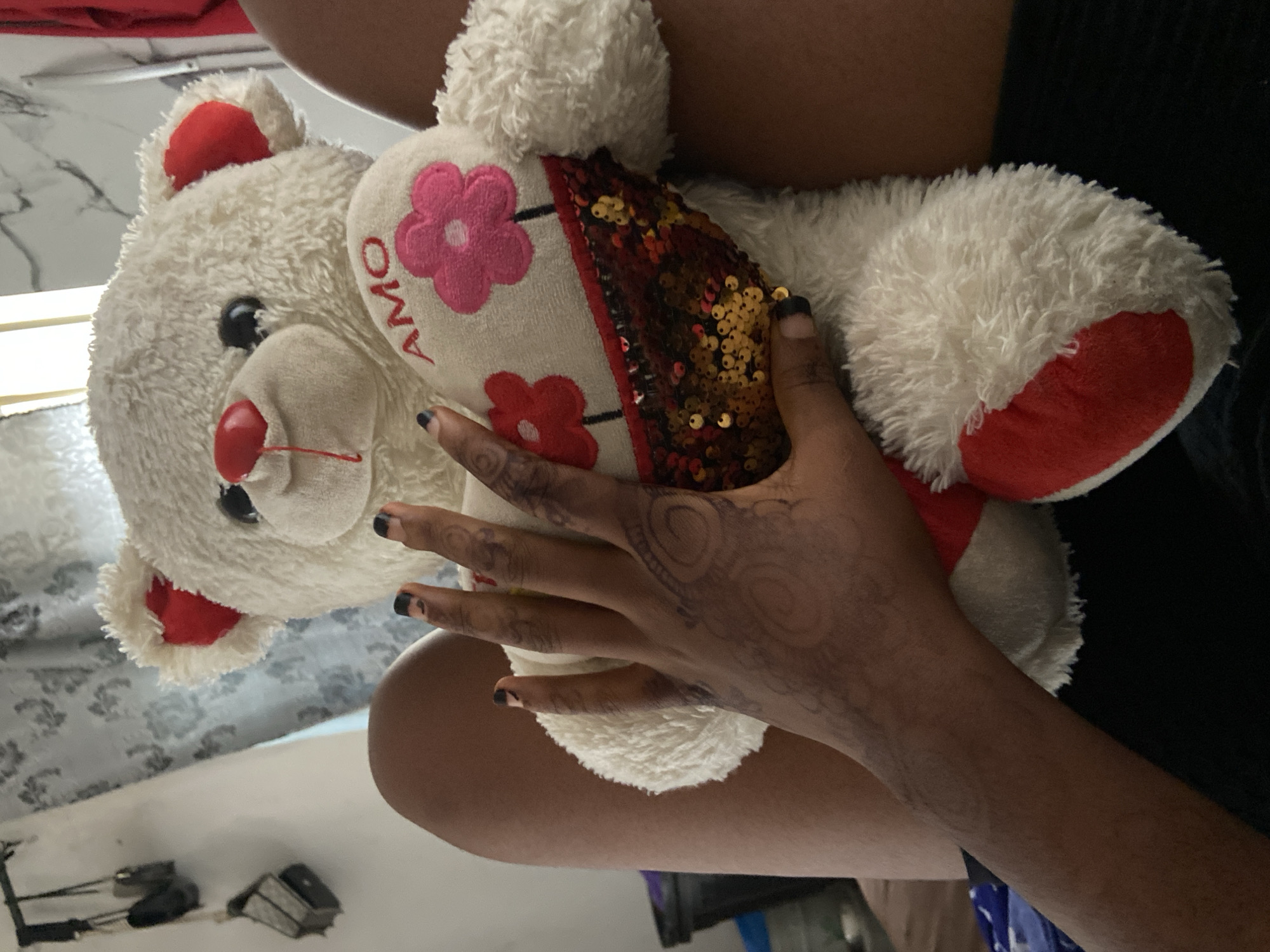 A person is holding a white teddy bear with red and pink accents. The teddy bear has a heart on its chest with the word "AMO" written on it. The person's hand has a henna design. The background includes a patterned surface, possibly a bed or floor, and some dark clothing. The location, Lokoja, Nigeria, is not discernible from the visual information.