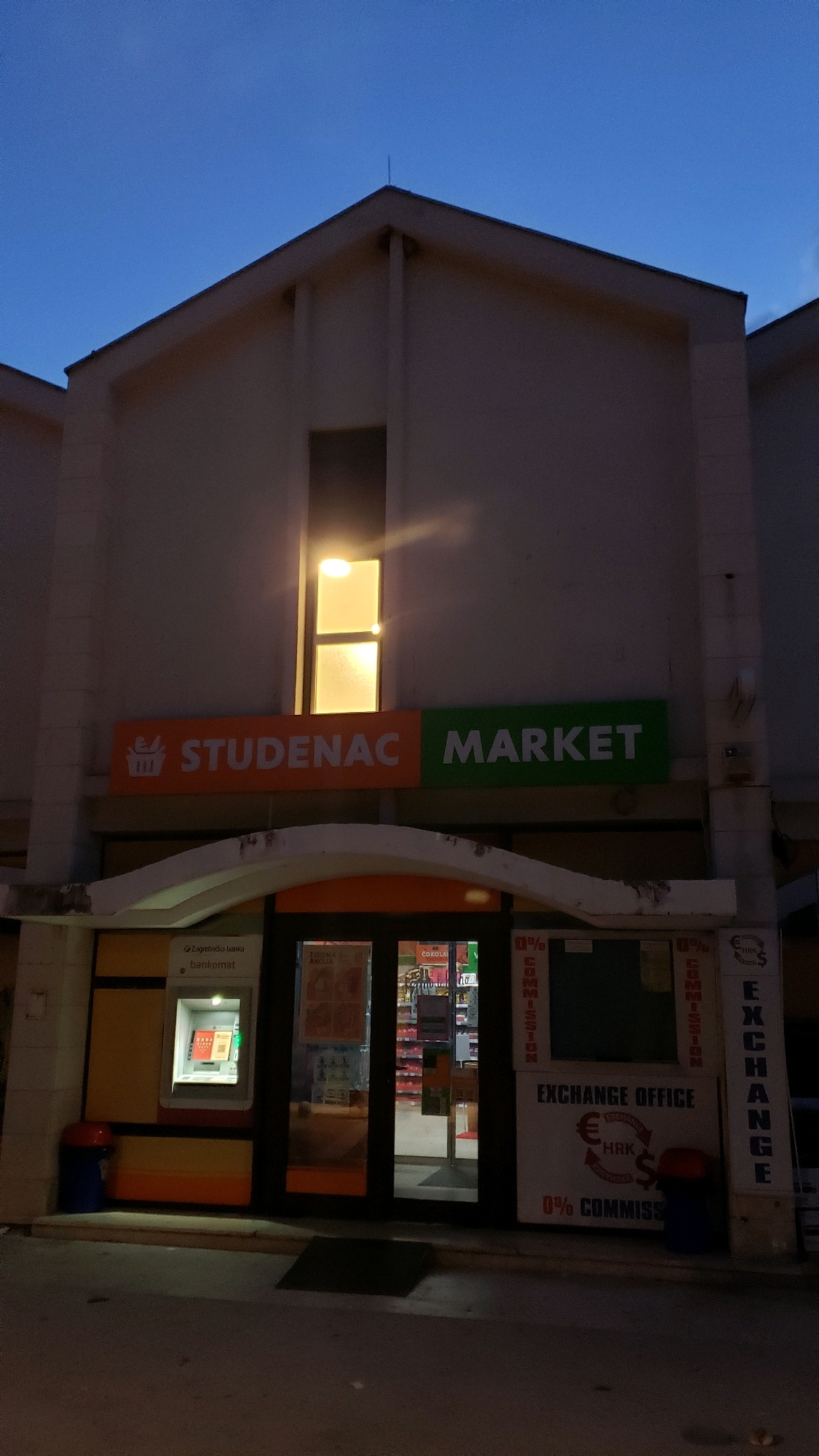The image shows the exterior of a Studenac market and an exchange office in Općina Tučepi, Croatia, at dusk. The building is two stories with a light-colored facade. A single rectangular window on the upper floor emits a warm yellow light. Below the window, a large sign with an orange background reads "STUDENAC" and a green background reads "MARKET". The entrance to the market is a double glass door. To the left of the entrance is an ATM with a "Zagrebačka banka" logo. To the right of the entrance is a sign for an "EXCHANGE OFFICE" with a circular diagram showing the exchange between Euros (represented by a euro symbol), Croatian Kuna (HRK), and Dollars ($), indicating "0% COMMISSION". The ground in front of the building is paved. There are two trash receptacles visible, one red and blue next to the ATM, and another blue one near the exchange office. The sky is a deep blue, indicating twilight.