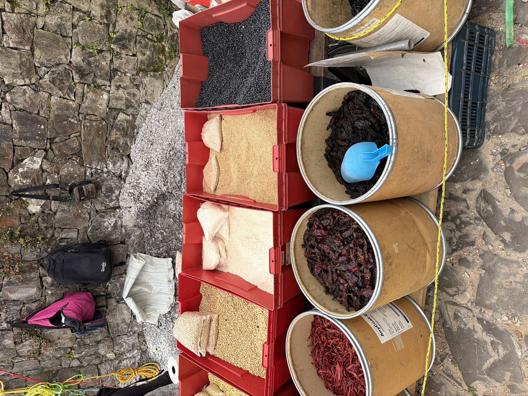 Here's a matter-of-fact description of the scene in Cuetzalan, Mexico:
The image shows a market setup on a stone surface, likely outdoors. On the right, stacked red plastic crates contain bulk dry goods including black beans, various grains, rice, and other beans. Next to them are cardboard barrels, each filled with different types of dried chili peppers. A blue scoop rests inside one of the barrels.
On the left side of the frame, several bags (including a backpack with the brand "Quala"), ropes, and some cloth are scattered on a stone surface. A camera bag also rests on the surface. The overall scene depicts a market stall.