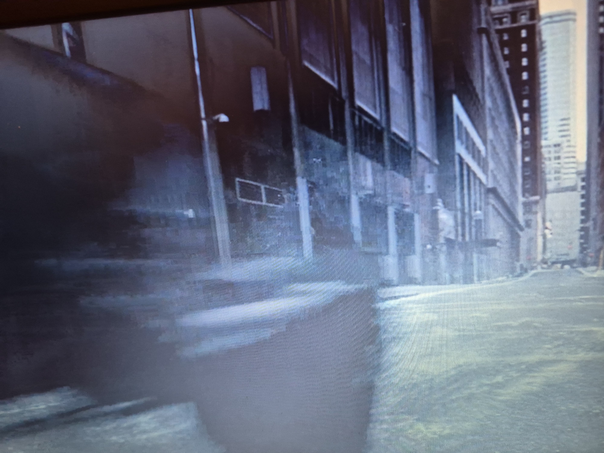 The image depicts an urban environment with a street scene. On the left, a dark, textured building with rectangular windows and a vertical light fixture is visible. To the right, a street stretches into the distance, lined by more buildings. The street itself appears wet or reflective, with subtle variations in color suggesting the presence of water or a glistening surface. In the background, taller buildings are visible, indicating a city setting. The overall atmosphere is somewhat dark and moody, with a focus on architectural elements and the urban landscape. There is no information in the image to suggest a location in Sandwell, United Kingdom.