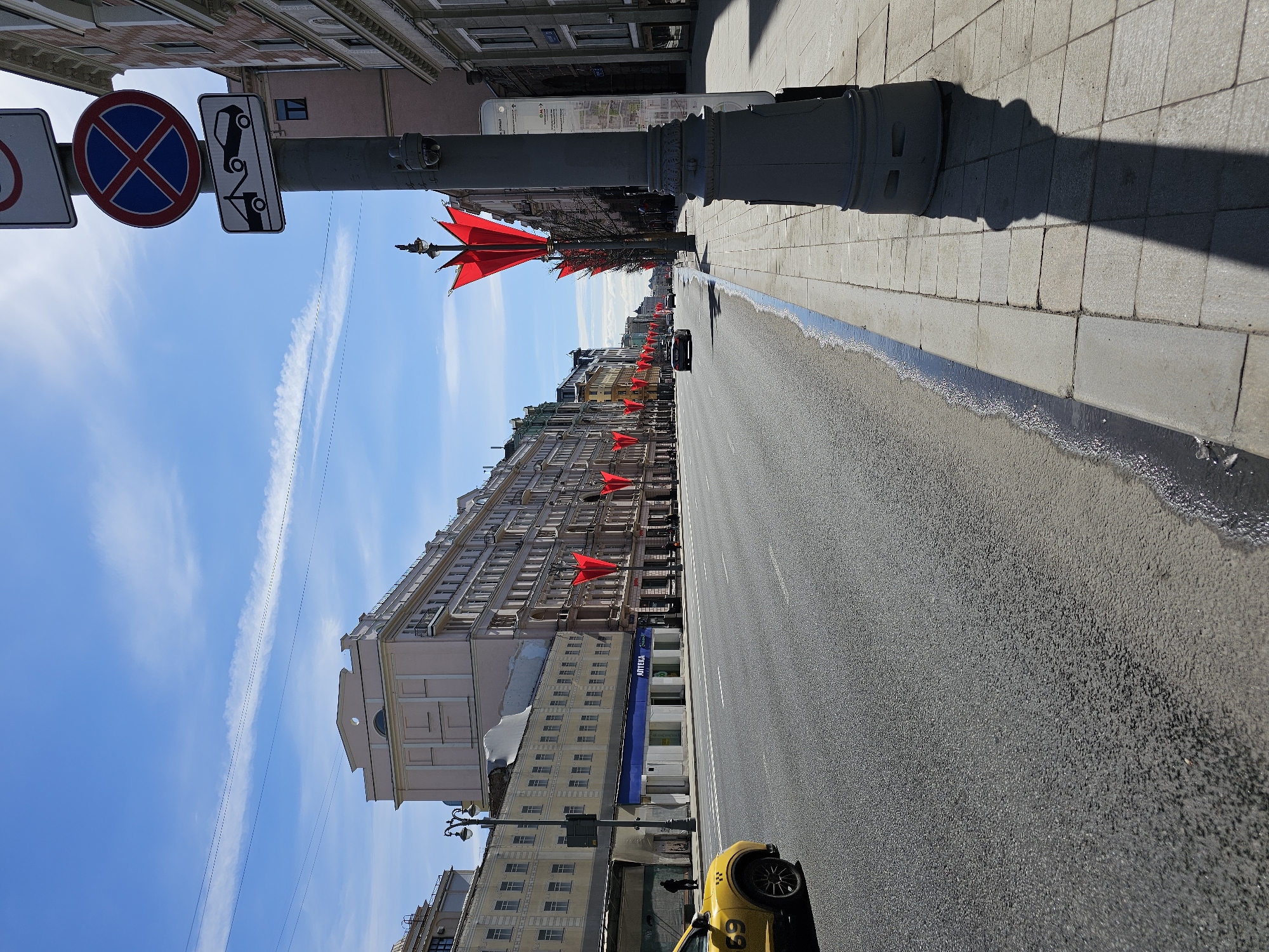 The image shows a street in Moscow, Russia. The street is lined with buildings, many with red flags hanging from them. A yellow taxi is parked on the right side of the street. The sky is blue with some clouds. Street signs are mounted on a pole in the foreground.