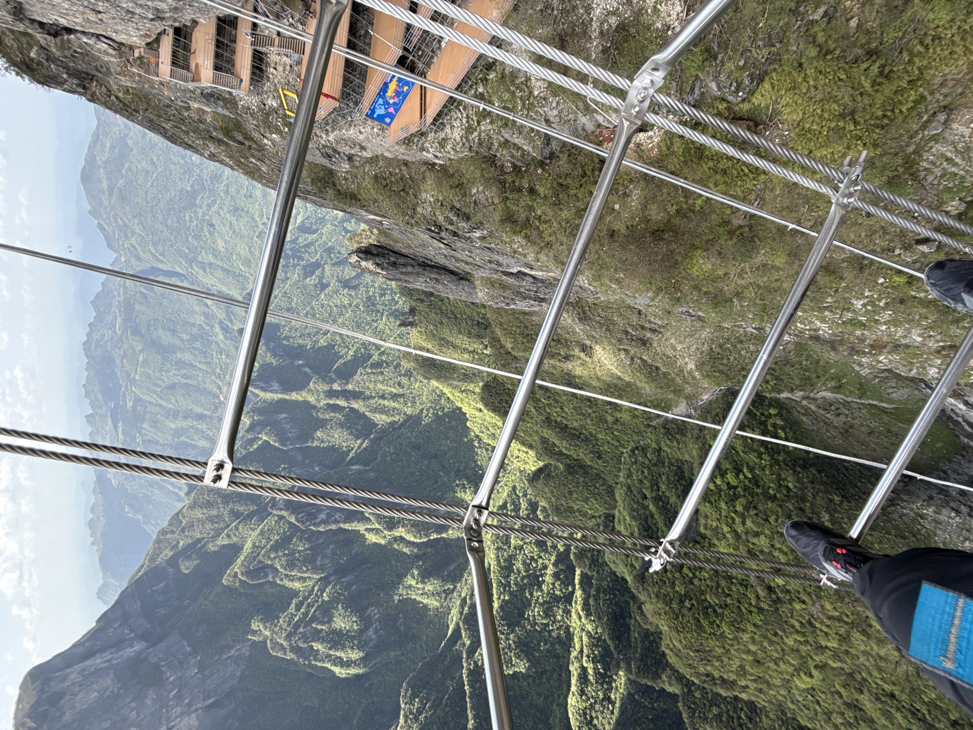 A low-angle, rotated shot shows a person standing on a metal and cable bridge system in a mountainous, verdant environment. The location is the city of Yongding District, China.
The person is wearing black pants and black athletic shoes with red accents. Their lower legs and feet are visible, indicating they are walking or standing on the bridge. The bridge consists of a metal framework with steel cable support.
The landscape below and to the side reveals a steep drop into a densely forested valley, and misty mountains. At the top left, part of a wooden platform with metal railings is visible.