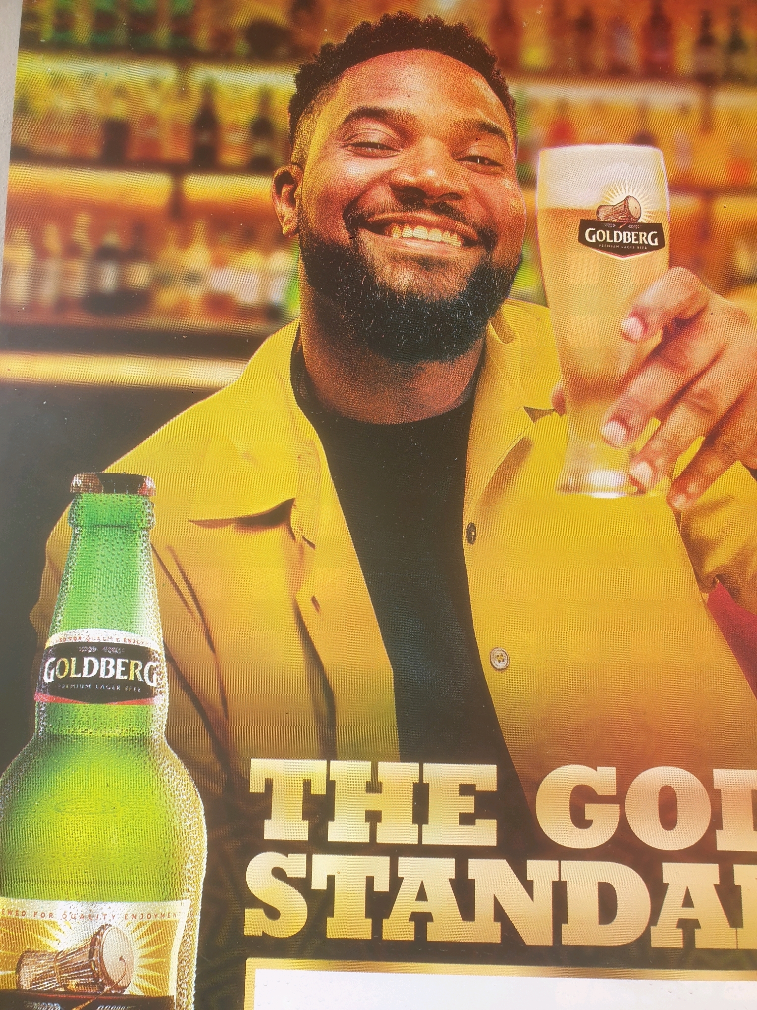 This image is a photograph of a man holding a glass of Goldberg beer and a bottle of Goldberg beer. The man is smiling and looking at the camera. He is wearing a yellow jacket and a black shirt. The background is blurred and appears to be a bar or a pub. The image also contains the text "THE GOLD STANDARD" which is likely a slogan for the beer. There is no indication in the image or the provided crops that this scene is located in Mangu Town, Nigeria.