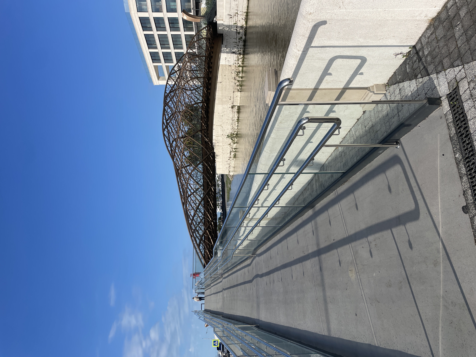 The image captures a section of a modern pedestrian bridge in Sochi, Russia, featuring glass and metal railings that cast long shadows on the concrete walkway. The bridge extends toward a unique architectural structure of weathered steel or iron, possibly a pedestrian bridge itself, positioned alongside a contemporary building with a grid of windows. Below, water flows beneath both bridges. The sky is clear and blue, with a few scattered clouds. Small figures of people are visible in the distance on the pedestrian bridge. A sign is also partially visible.