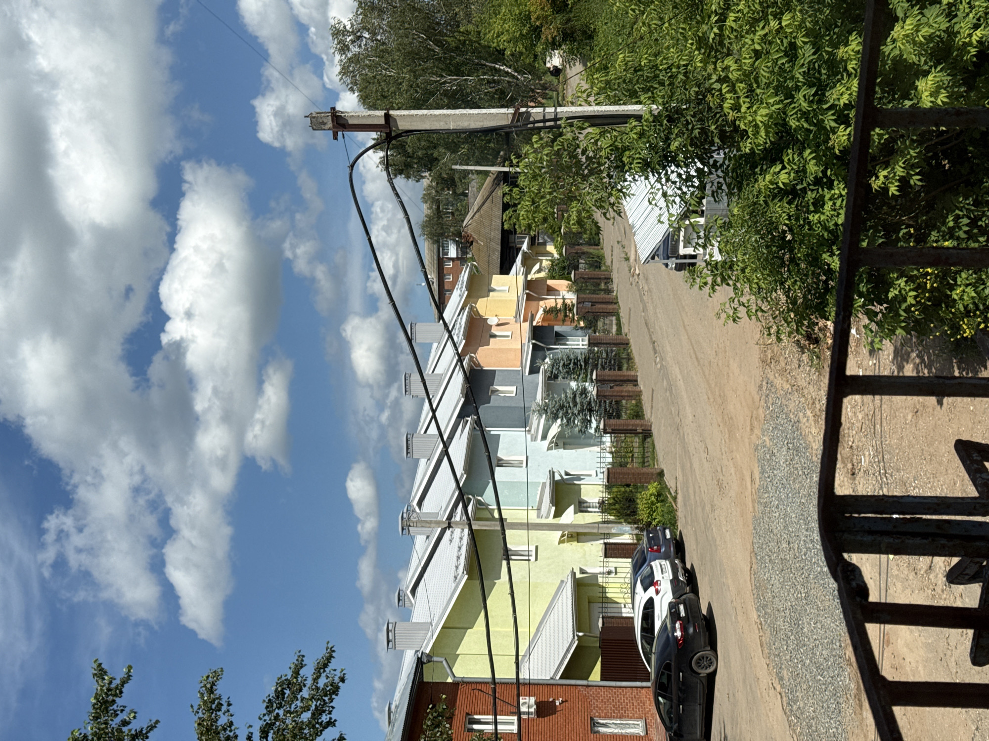 The image depicts a row of colorful townhouses in Votkinsk, Russia, under a bright blue sky with scattered white clouds. The townhouses, painted in shades of brick red, lime green, light blue, light orange, and yellow, are arranged along a dirt road. Each house has a light-colored, sloped roof with multiple chimneys. A black metal fence and gate are visible in the foreground, with a gravel area in front of it. Several cars, including a black sedan, a white SUV, and a dark blue sedan, are parked along the side of the road in front of the houses. Electrical wires crisscross the scene, supported by a concrete utility pole. Lush green trees and bushes frame the right side of the image, and more trees are visible in the background. The scene appears to be a residential street.