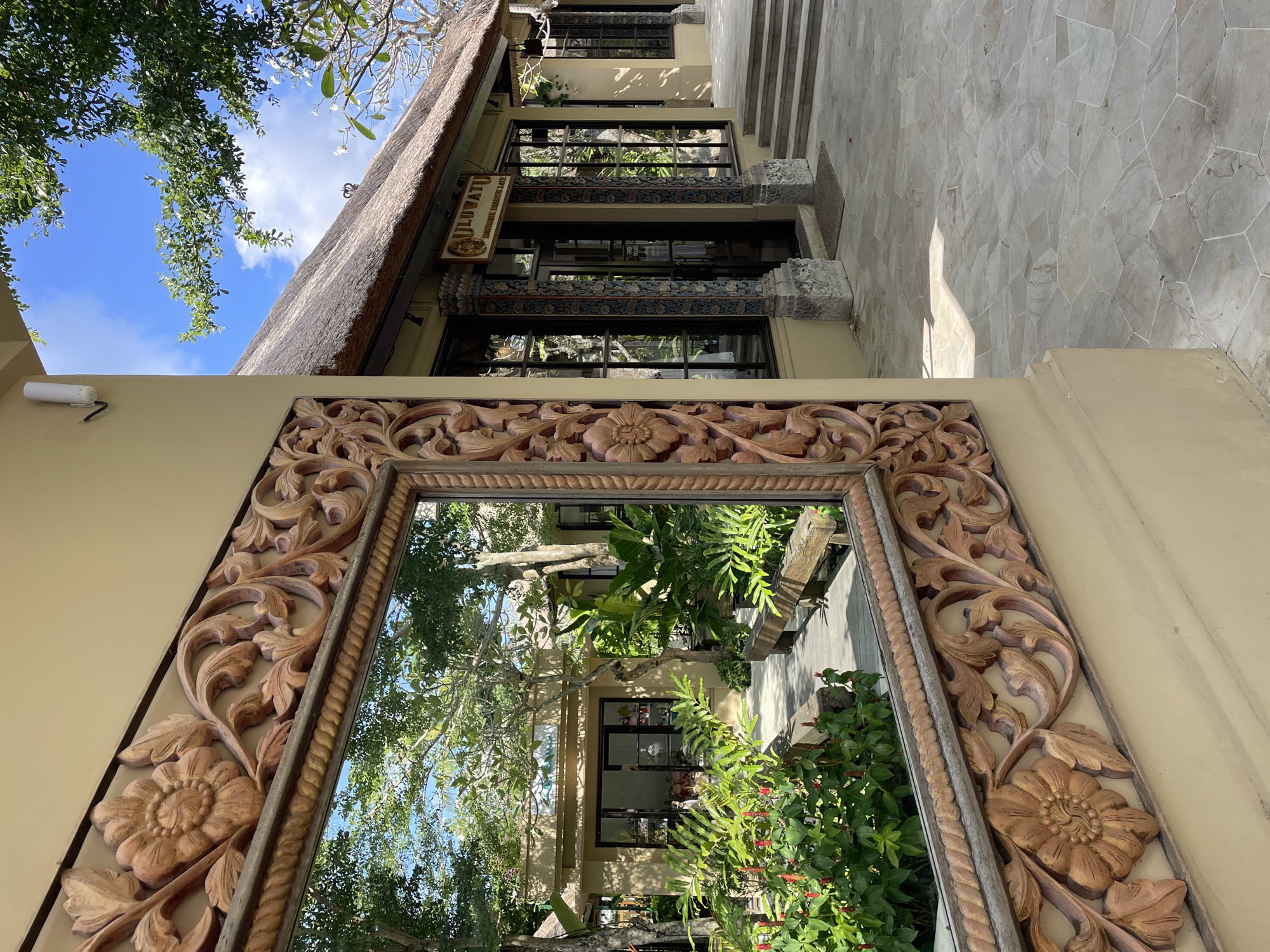 The image captures a mirror with an ornate wooden frame, reflecting an outdoor scene of a building with a thatched roof and the signage "JILUVATU Boutique Marine Art." The mirror's frame features carved floral and leaf designs. The reflection reveals vegetation, including trees and flowering plants, in front of the building. Above the building, a clear blue sky is visible with light clouds and tree branches. The location is in Unknown, Unknown.