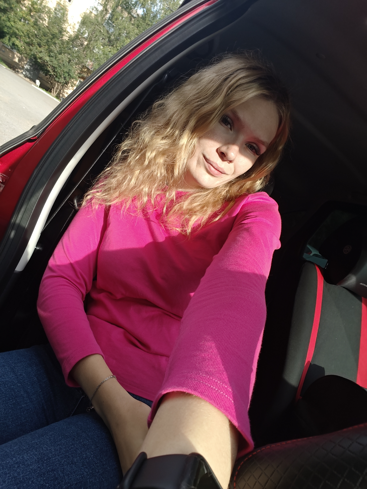 An outdoor, close-up, selfie-style photograph shows a young, Caucasian woman with blonde, wavy hair and light skin, wearing a bright pink long-sleeved shirt and blue jeans. She is seated in the passenger seat of a car, with the car door open. The interior of the car appears to be red and black. The woman is looking at the camera and smiling slightly. The background of the image shows trees and a building, suggesting an urban or suburban environment. The sunlight casts shadows on the woman's face and clothing. The location is the city of Izhevsk, Russia, specifically a street in the city.