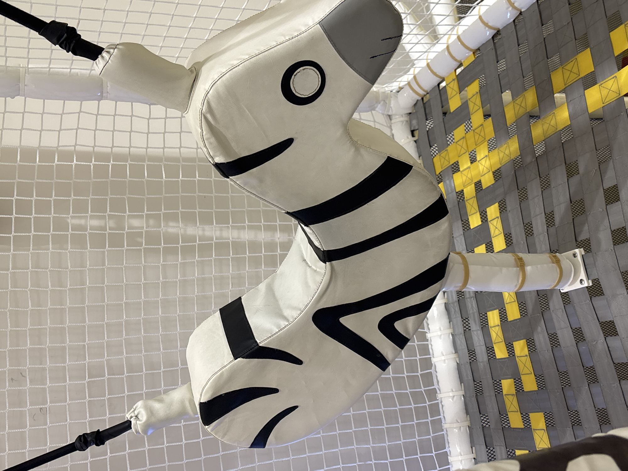 This is a close-up, eye-level, full-shot photograph of a stuffed zebra-themed ride-on toy hanging from a white net enclosure. The toy is primarily white with black stripes, mimicking a zebra's pattern. It is suspended by black ropes attached to white padded supports. The background consists of the white net enclosure and a grey and yellow woven floor. There is no visible signage or identifiable landmarks indicating a specific location in Dubai, United Arab Emirates. No people are present in the image.