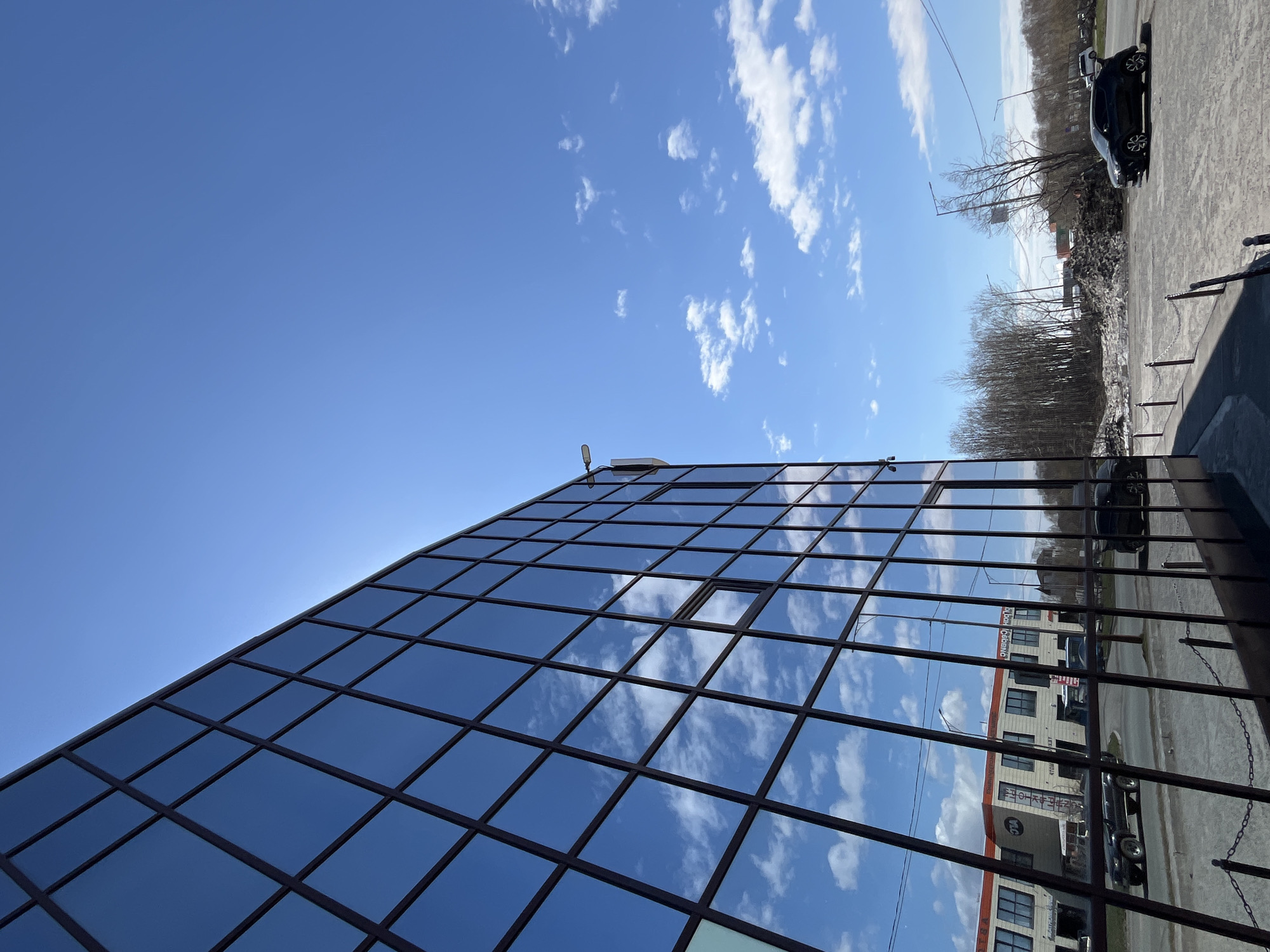 The image shows a building with a glass facade divided into square panes. The facade reflects the sky and clouds. One window is open. A car is parked on the street in front of the building. The building in the reflection appears to be a commercial structure. Bare trees and remnants of snow are visible. The location is identified as Petropavlovsk-Kamchatsky, Russia.