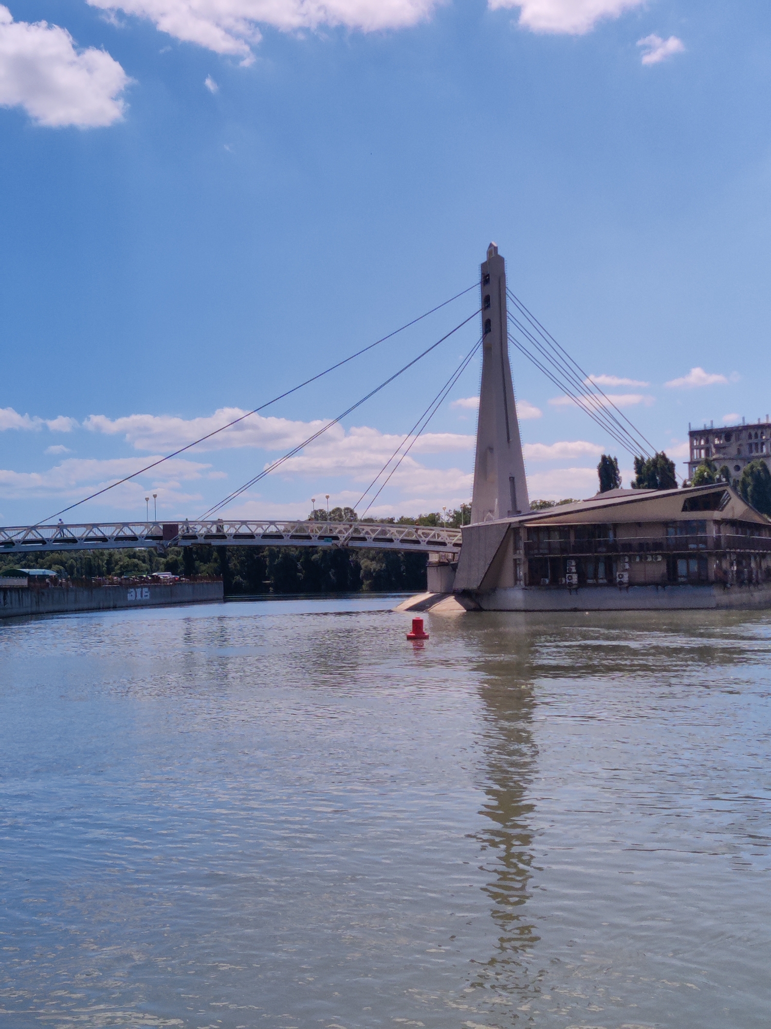 The image shows a wide river with a cable-stayed bridge crossing it. On the right side of the river, a modern building, possibly a restaurant or event venue, is situated near the bridge's support tower. The building has multiple windows and balconies. In the background, more buildings and lush green trees can be seen along the riverbank. The sky is blue with scattered white clouds. A red buoy floats in the river in the foreground. The bridge itself is a suspension bridge with a tall central tower and cables supporting the roadway. Part of a barge with graffiti is visible on the left side of the river. This scene is located on the Kuban River in the city of Krasnodar, Russia.