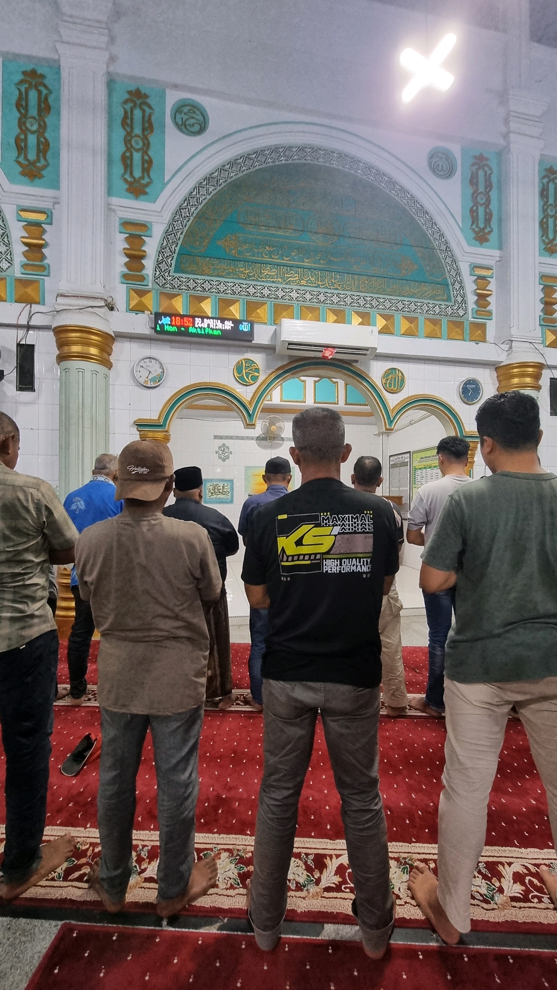 In the city of Puloara, Indonesia, a group of men are engaged in congregational prayer within a mosque. The men are positioned on a red carpet, facing towards the front of the mosque, which features an arched mihrab decorated with Islamic calligraphy and geometric patterns in turquoise and gold. The walls of the mosque are white with ornate gold and turquoise accents. Above the mihrab, a bright, cross-shaped light fixture illuminates the space. In the background, a digital display shows the time as 18:52, along with prayer schedule information. Several men are seen from behind, wearing prayer attire including a black t-shirt with graphics, a brown t-shirt, a blue shirt, and light-colored trousers. Some are barefoot, while others wear shoes. The overall atmosphere suggests a moment of religious observance.