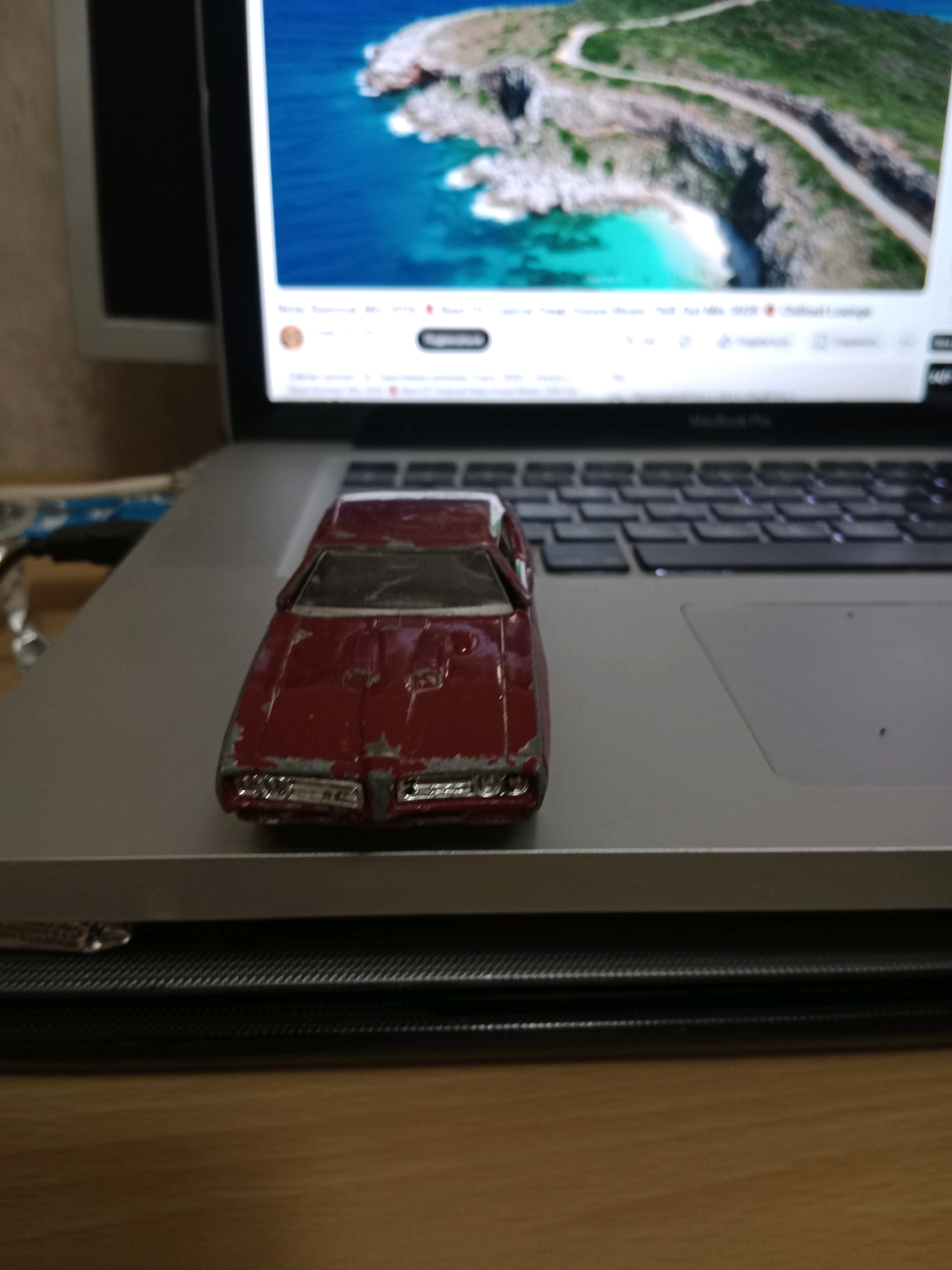 A red die-cast toy car, exhibiting wear and tear, rests on a silver laptop. The laptop's screen displays a scenic landscape with a turquoise ocean and a winding road atop a cliff. The laptop is situated on a dark, textured surface, which is placed on a wooden table. The overall scene is a close-up shot of the toy car and laptop, with the background out of focus. There is no indication of the location being in Bălăbănești, Moldova.