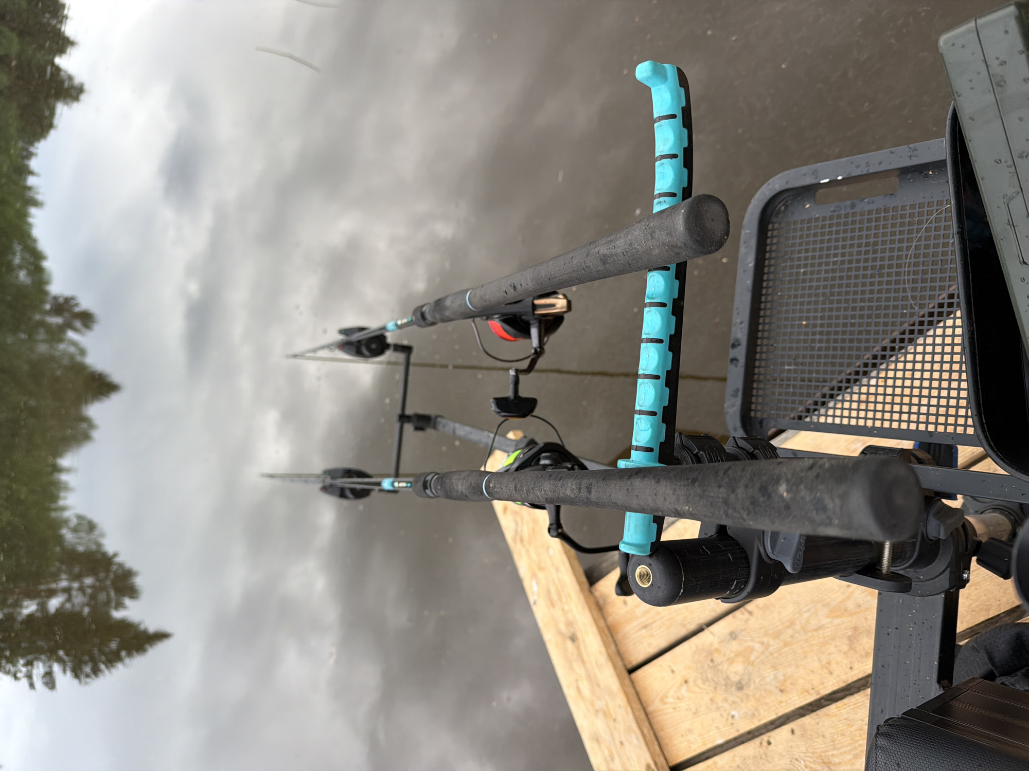 The image depicts two fishing rods mounted on a support structure, likely part of a fishing platform or chair. The rods are dark gray, with fishing reels attached. One rod has a bright blue support piece with a laddered design. The support structure is a dark gray, metallic frame with a mesh seat. The rods are positioned over water, which reflects the cloudy sky and trees on the opposite bank. The foreground shows the wooden planks of the fishing platform or dock. The exact location within Прикамский, Russia cannot be determined with the provided information. There are no people visible in the image.