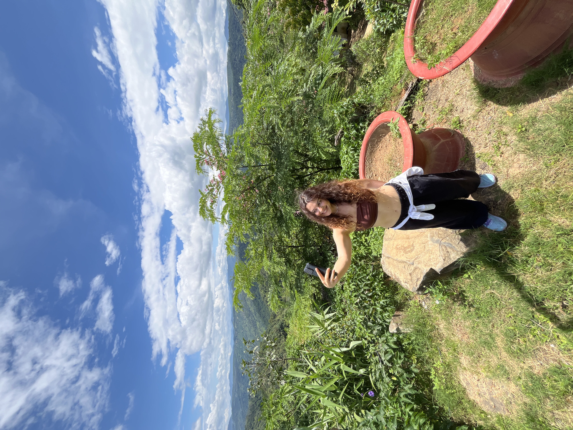A young woman with curly brown hair and a brown tube top is taking a selfie with her phone. She is standing on a grassy area next to a large rock and two large, red planters. She is wearing black pants with a white shirt tied around her waist and blue sneakers. The background is a lush green landscape with rolling hills and a bright blue sky with white clouds. The scene is outdoors, possibly at a viewpoint in Sơn Điền, Vietnam, given the verdant, hilly terrain and tropical vegetation.