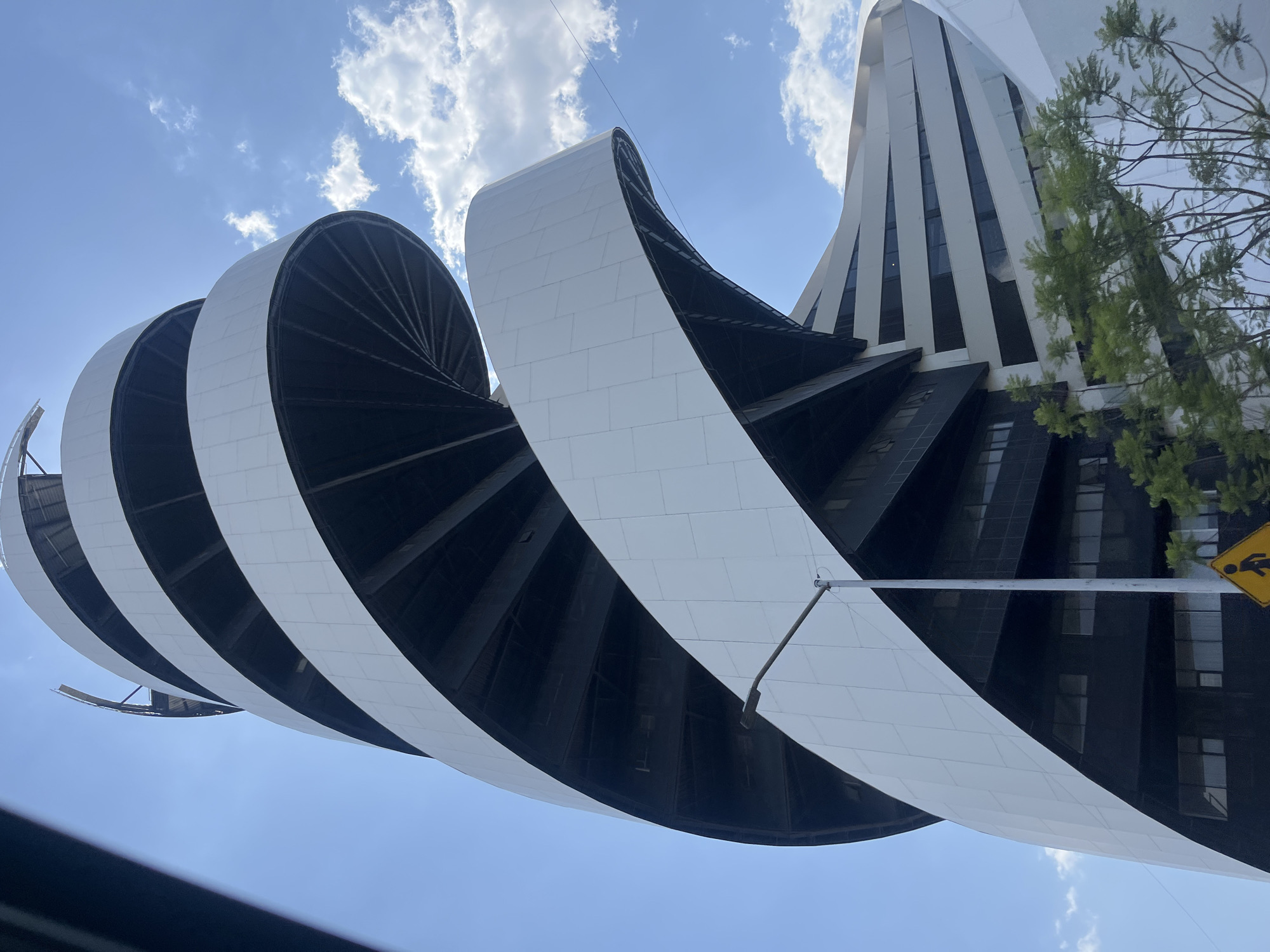 The image shows a low-angle view of the Museo Internacional Barroco (International Baroque Museum) in San Bernardino Tlaxcalancingo, Mexico. The building features a distinctive curvilinear design with a spiral structure. The spiral consists of alternating black and white segments. The white sections appear to be constructed of rectangular panels or tiles. The museum's main building can be seen in the background, with white vertical fins. A blue sky with scattered clouds is visible. Parts of a tree and a yellow pedestrian crossing sign are also in the background.