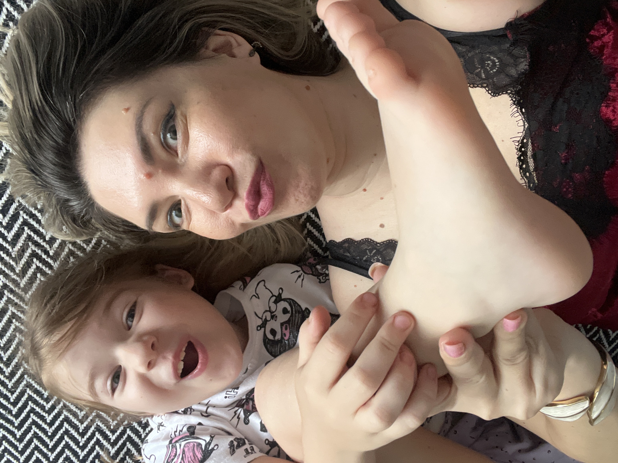 A woman and a young girl are lying down, looking up at the camera. The girl is in the foreground on the left, with her mouth open in a wide smile. She is wearing a white t-shirt with black illustrations of animals. The woman is to the right, with her eyes wide and her lips pursed. She is wearing a black lace top. The woman is holding the girl's foot, which is in the center of the image. The background is a black and white patterned blanket. The scene appears to be indoors, likely a bedroom or living room. There is no indication of the city of Sochi, Russia.
