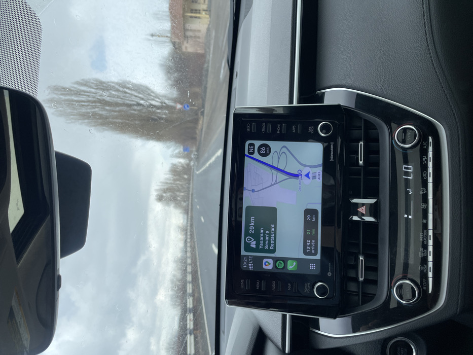 The image captures an interior view from a moving vehicle on a highway in Solak, Armenia, at approximately 13:21 under an overcast sky with visible moisture on the windshield.
The foreground is dominated by the vehicle's black dashboard and central infotainment system. The infotainment screen, displaying a navigation application (likely Apple CarPlay), shows the current time as 13:21 and an estimated arrival time of 13:42, indicating a 21-minute journey over 29 kilometers to "Yasaman Sevan's Restaurant." The vehicle's current speed is registered at 86 km/h, heading northeast (NE) on a road labeled "AH83." Below the screen, the vehicle's climate control panel indicates a temperature setting of "LO" with "SYNC" active. A red triangular hazard light button is centrally located between air vents.
Through the windshield, a multi-lane asphalt highway is visible, curving slightly to the right. A white corrugated barrier runs along the left side of the road. The right side of the highway is lined with tall, slender, bare deciduous trees. Further beyond the trees, a small, light-colored, single-story building with a dark roof is visible. Several blue road signs are partially visible on the right shoulder, beyond the trees. The sky is uniformly grey with heavy cloud cover, and small water droplets are evident on the windshield's exterior surface, suggesting rain or recent precipitation. No individuals are visible in the scene.
