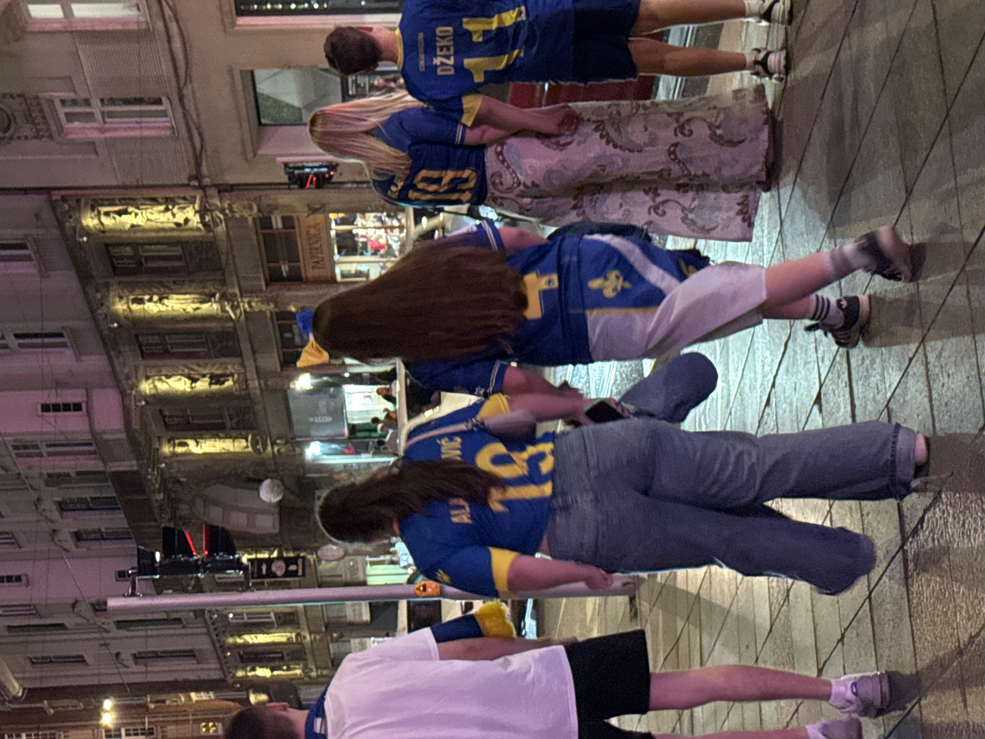 This image depicts a street scene at night in Sarajevo, Bosnia and Herzegovina. The primary subjects are a group of young people wearing matching blue and yellow football jerseys. In the foreground, from left to right, is a person in a white t-shirt and black shorts. Next to them is a young woman wearing a blue and yellow jersey with the name "ALA JOVIĆ" and the number "19" on the back, paired with light blue jeans. Beside her is another young woman in a similar blue and yellow jersey, with a decorative patterned skirt. On the far right is a young man also wearing a blue and yellow jersey, with the name "DŽEKO" and the number "11" on the back. They are walking on a wet, tiled sidewalk, reflecting the city lights. The background shows a street with buildings exhibiting distinct architectural styles, some illuminated with yellow lights, and a traffic light visible. The scene suggests a gathering or outing, possibly related to a sporting event.