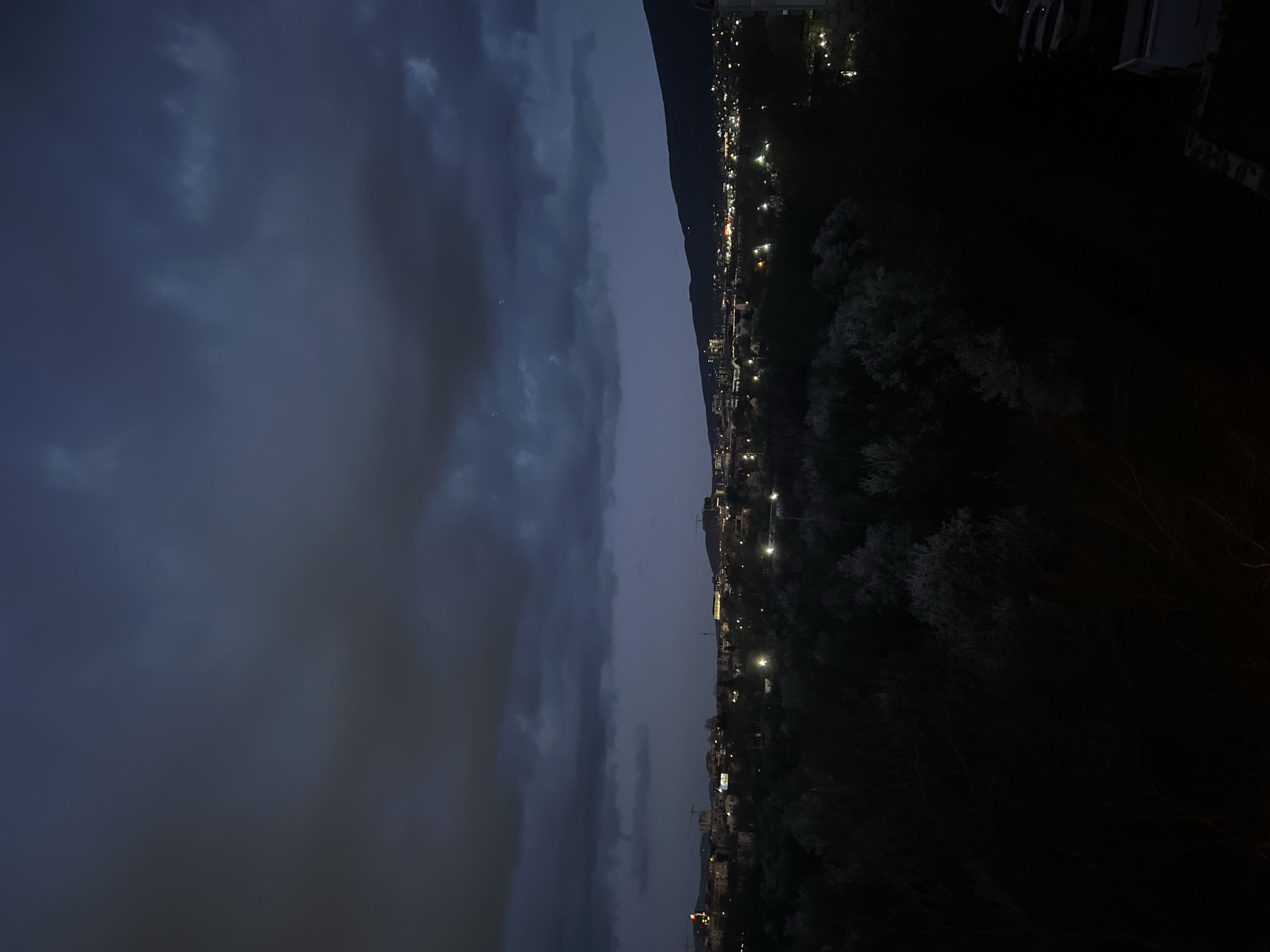 The image depicts a nighttime view of a city from an elevated position. On the left side, there is a dark, cloudy sky. In the middle, a dark mountain or hill range is visible. On the right, lights from the city illuminate buildings, streets, and trees. The scene is likely in the city of Sofia, Bulgaria, but without further context, precise location identification within the city is not possible. The foreground is mostly dark, suggesting a natural or undeveloped area.