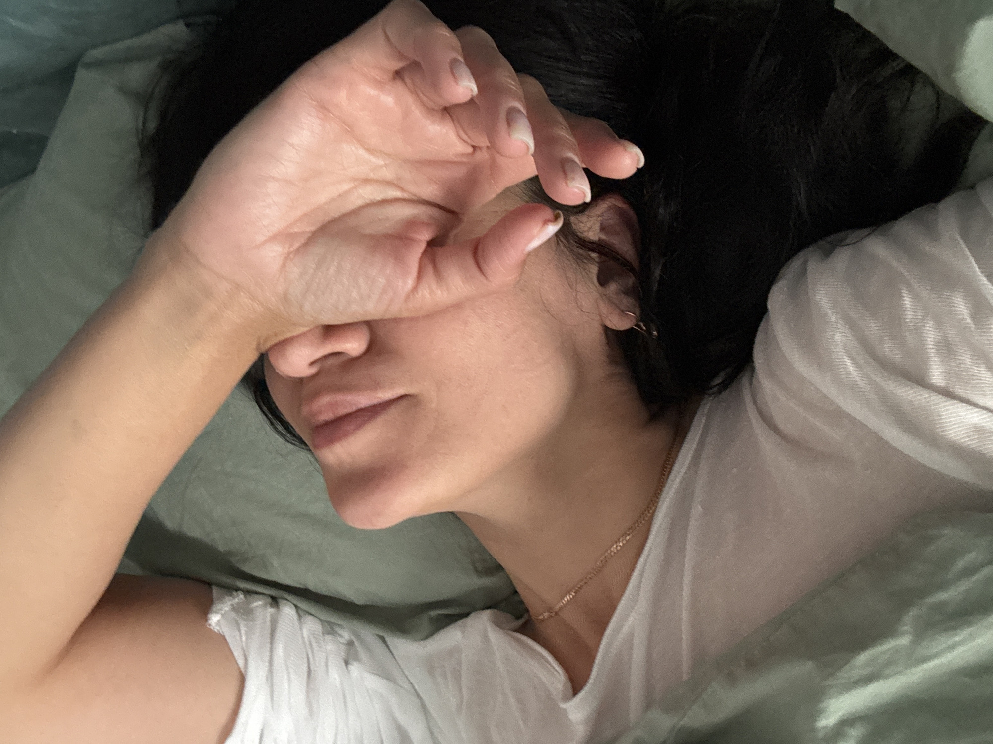 A person's face is partially obscured by their hand as they lie down on a bed with light green sheets. The person is wearing a white t-shirt and a thin gold chain necklace. Their dark hair is visible around their head. The image captures a close-up view of the person's face, neck, and arm, with the rest of the scene out of focus. There is no discernible information about the city of Chaikovsky, Russia, or any other specific location within the image.