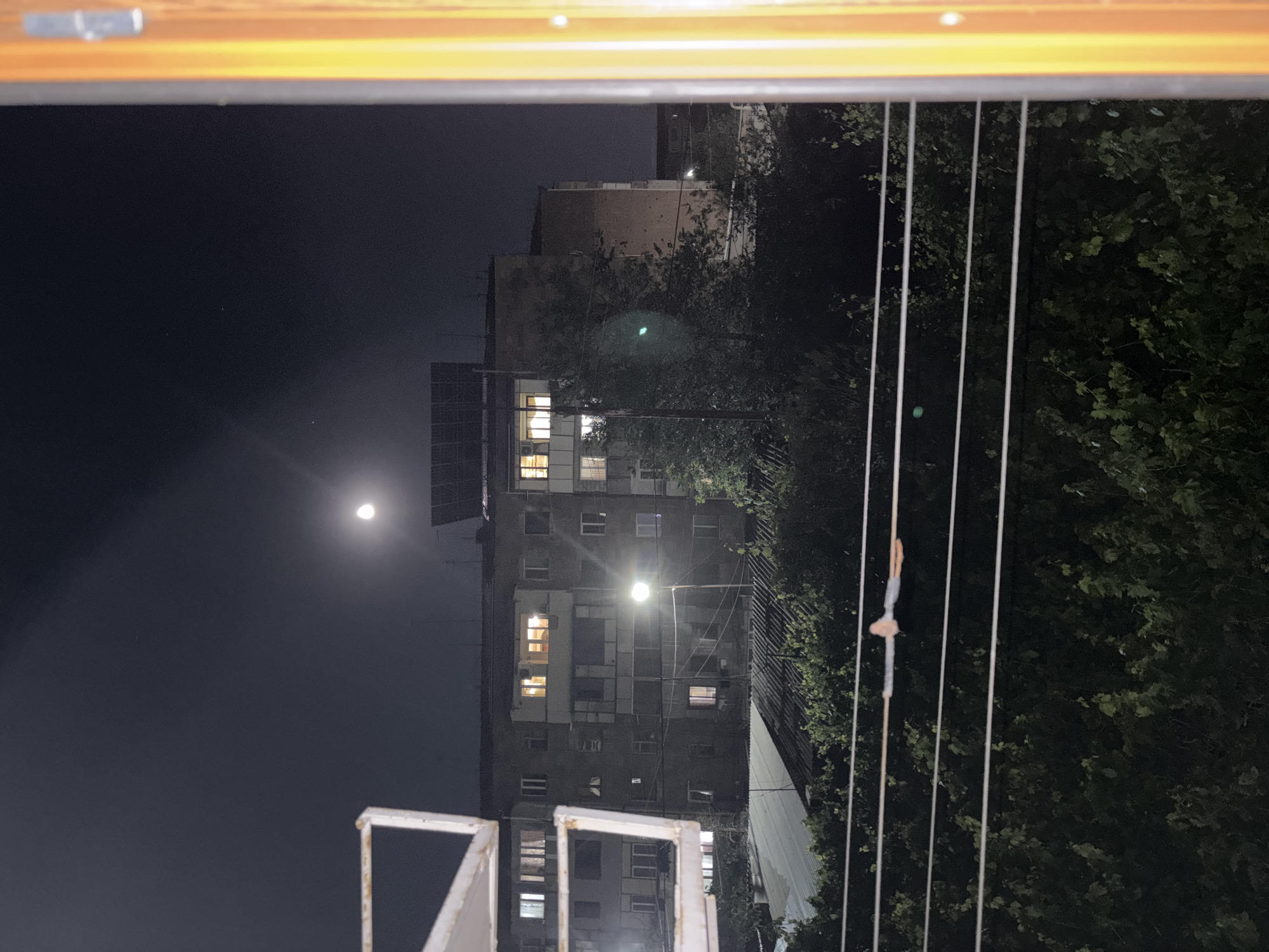 View from a window at night, possibly in Vagharshapat, Armenia. A multi-story building with illuminated windows is visible. The sky is dark with a visible crescent moon. Trees and power lines are also in the view.