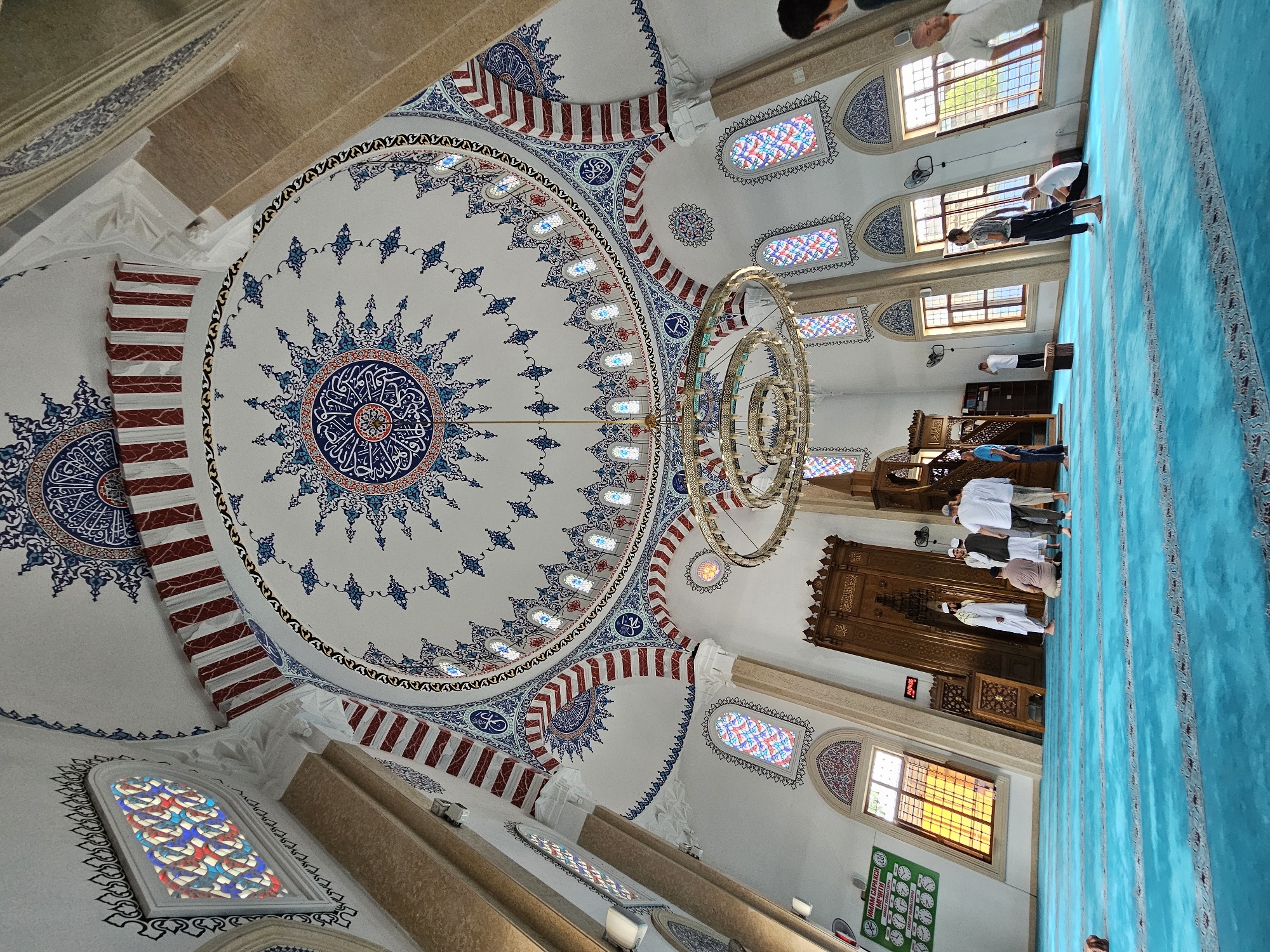 This is an interior shot of a mosque in the city of Özgön, Kyrgyzstan. The mosque features a large, ornate dome decorated with intricate blue and white Islamic calligraphy and geometric patterns, as well as red and white striped arches and stained-glass windows. A large circular chandelier hangs from the center of the dome. The floor is covered with a bright blue carpet with gray patterned borders. Several men are present in the mosque, some standing and some kneeling, appearing to be engaged in prayer or conversation. The men are dressed in traditional Islamic attire, including white robes and caps. A wooden mihrab and minbar are visible against the back wall. The overall atmosphere is serene and spiritual.