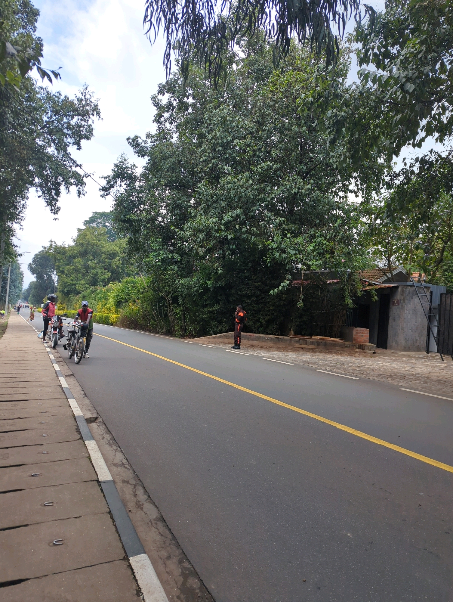 The image shows a paved road with a yellow line dividing the lanes, likely in Musanze District, Rwanda. A concrete sidewalk runs along the left side of the road. Several people are present. On the sidewalk, a person wearing a red jacket and helmet stands next to a motorcycle. Another person, wearing a red shirt and a cap, is pushing a bicycle on the sidewalk. Further down the road, a person in an orange and black uniform is standing near a building. The area is surrounded by lush green trees. A building with a dark door and a ladder on its side is visible to the right. The sky is cloudy.