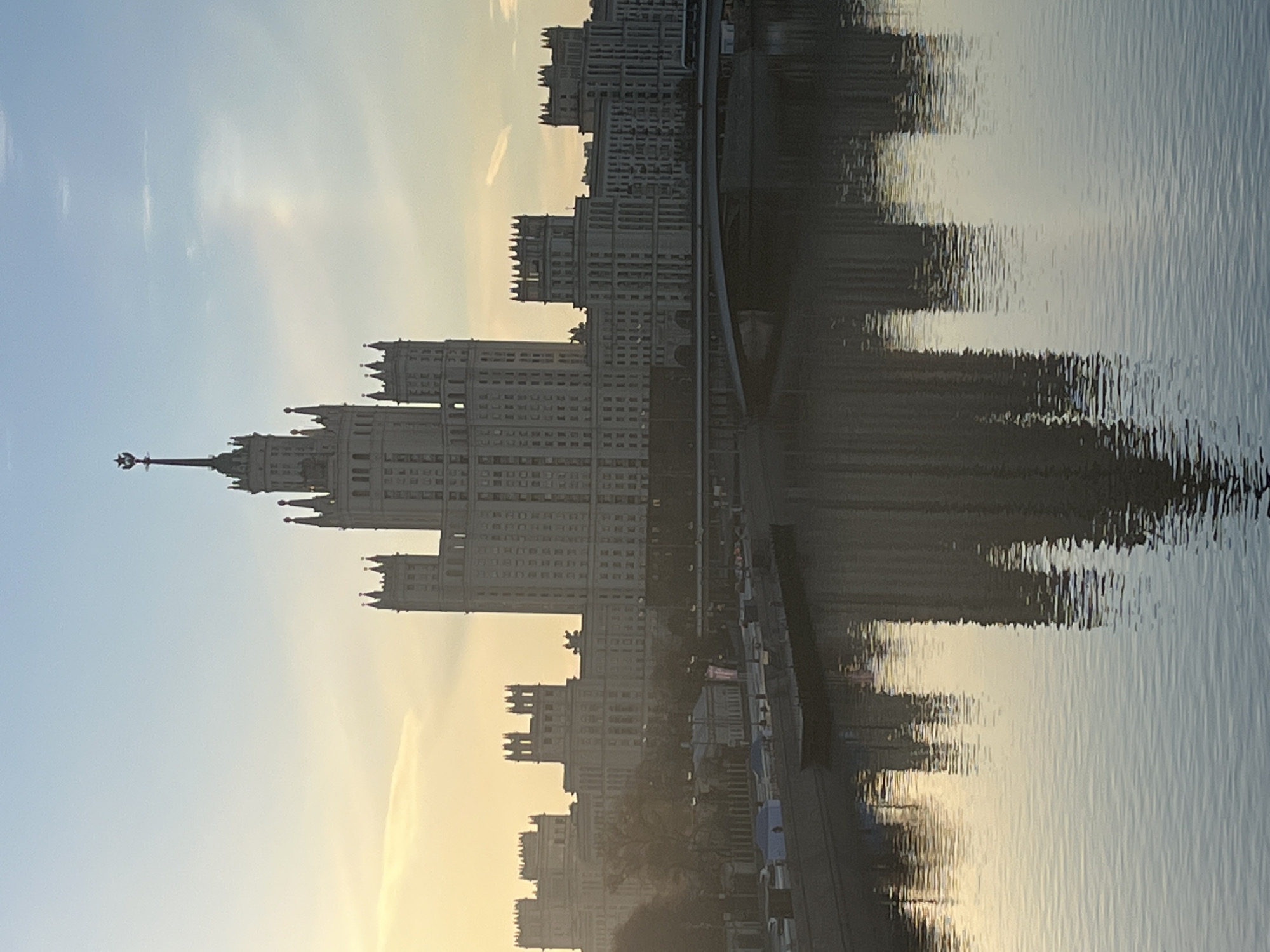 The image shows a building reflected in water. The building is one of the Seven Sisters, a group of skyscrapers in Moscow designed in the Stalinist style. The water is likely the Moskva River. The sky is light blue with some clouds. It is daytime.