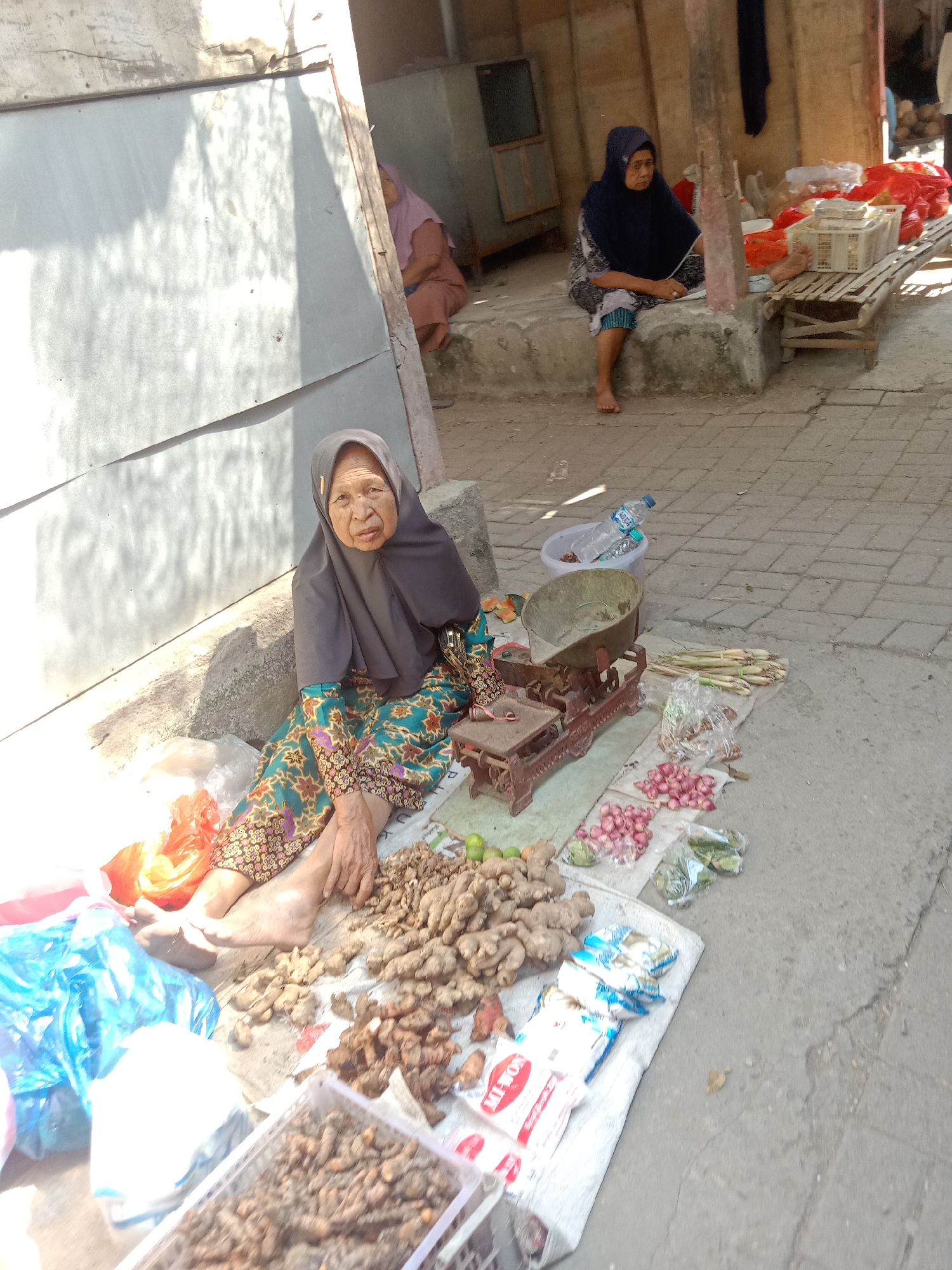 In Klepek, Indonesia, an elderly woman wearing a gray hijab and a patterned green dress sits on the ground next to a collection of produce. Her legs are crossed, and her bare feet are visible. She is positioned in front of a stall with various items laid out on the pavement. In front of her is a vintage red weighing scale with a metal bowl. Scattered around the scale are onions, lemongrass, and what appears to be ginger and turmeric roots. Several plastic bags, some blue and some orange, are also near her. In the background, another woman in a dark hijab is seated on a raised platform, with baskets of goods behind her. The scene appears to be an outdoor market.