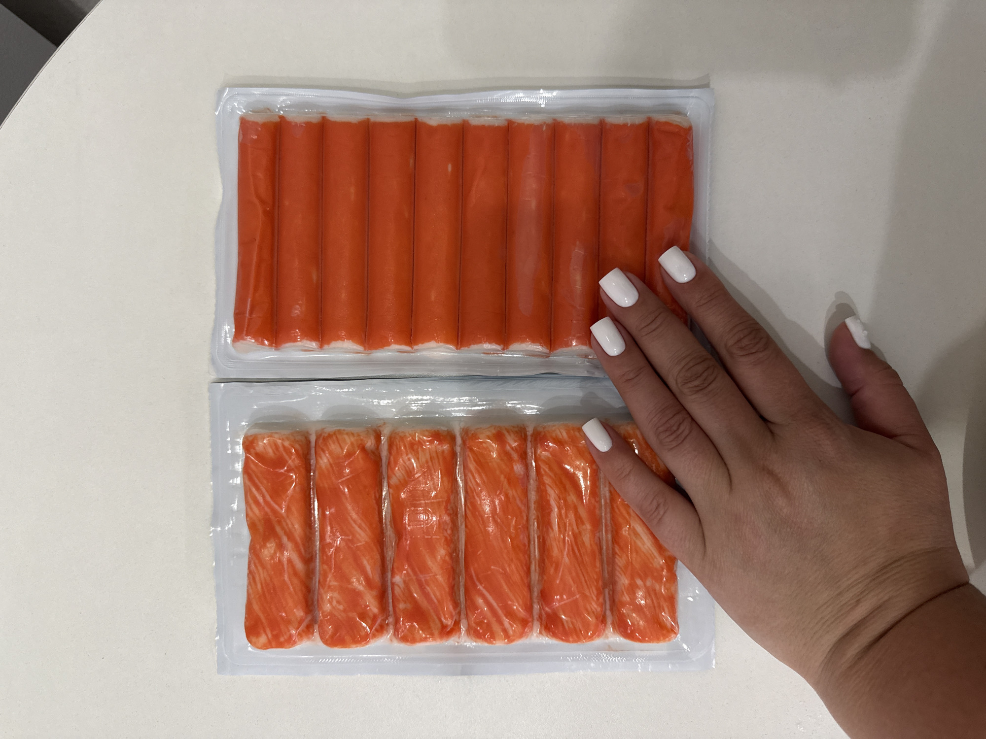 In Vologda, Russia, on a light-colored countertop, two plastic packages of imitation crab sticks are displayed side-by-side. The package on the left contains six sticks with a white and orange striated appearance, characteristic of imitation crab. The package on the right holds eight uniformly orange sticks, suggesting a different preparation or flavoring. A woman's hand, adorned with white nail polish, rests on the packages, her fingers gently touching the right side of the left package and the left side of the right package, indicating interaction with the products. The background is a neutral, light-colored surface, likely a kitchen counter, with a subtle shadow on the right side suggesting lighting from above and to the left.