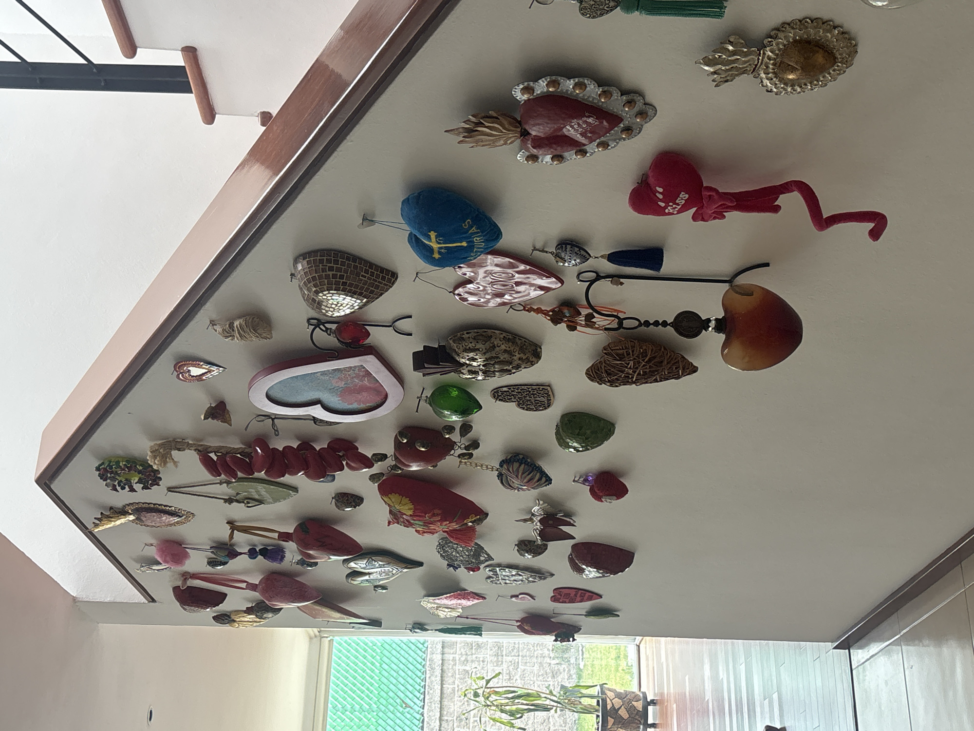 The image depicts a collection of decorative objects affixed to a ceiling, likely in a private residence. The collection includes a variety of items, such as hearts, religious symbols, ornamental tassels, beads, and stones. The objects are of varying sizes, shapes, and materials, suggesting a diverse collection accumulated over time. In the background, a window provides a view of greenery, possibly a garden. Considering the style and subject matter of the objects, specifically the prevalence of hearts and religious symbols, it is possible this is a private residence in Puebla, Mexico. No people or animals are visible in the image. The objects are static, attached to the ceiling. The environment is interior.