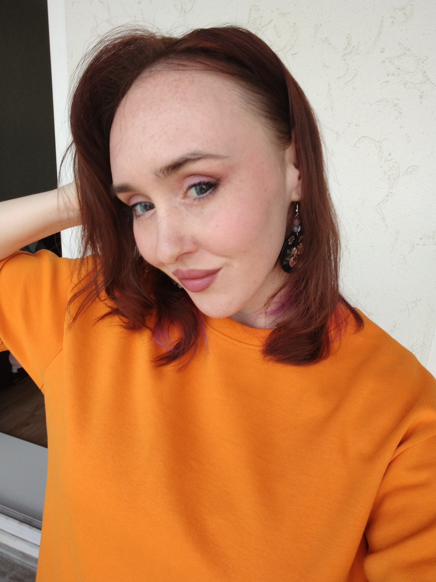 A close-up selfie or portrait shows a woman with reddish-brown hair, with subtle purple streaks visible near the ends. She has light skin with freckles across her forehead and nose. Her light-colored eyes are made up with pink eyeshadow, and she wears plum or mauve lipstick. She is dressed in a bright orange long-sleeved top or sweatshirt. On her right ear (viewer's left), she sports a dark, tear-drop shaped earring adorned with floral or abstract patterns and small beads.
The woman is looking towards the camera with a gentle, slight smile, her head tilted to her right. Her left arm is raised and positioned behind her head or neck, contributing to her pose. The background features a light-colored, textured wall, suggesting an indoor setting or a covered outdoor space like a balcony in Izhevsk, Russia. The lighting is bright and even, indicating it is daytime. There is no visible text in the image.