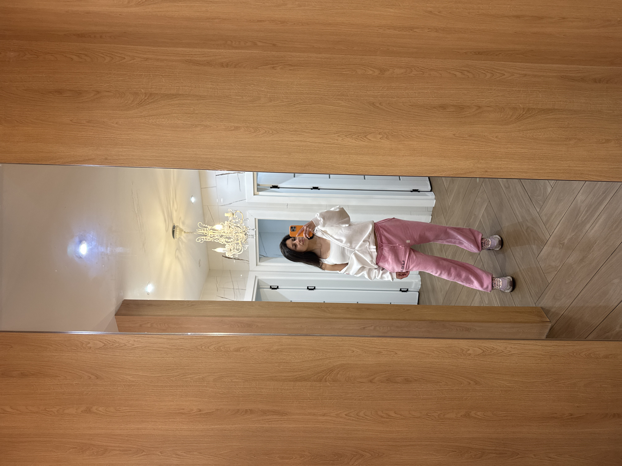 A mirror reflects a scene of a young woman taking a selfie in what appears to be the interior of a residence. The woman is wearing pink sweatpants, a white top, and sneakers. She's holding a smartphone up to take the picture. The background reveals a hallway with multiple white doors and a chandelier. The floor is a light brown herringbone pattern. The frame of the mirror itself has a light wood grain pattern. Without additional information, determining the exact location in Blagoevgrad, Bulgaria, is not possible.