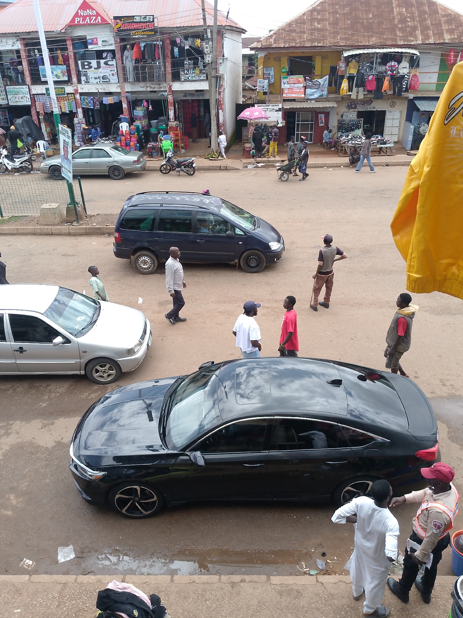 This image captures a street scene in Bukuru, Nigeria, likely in a commercial area with multiple shops. In the foreground, a black sedan is parked on a wet, muddy surface, with its hood reflecting the sky. To the left, a silver sedan is parked. A dark blue minivan is visible further back, to the right of the silver sedan.
Several individuals are present in the scene. Two men in the foreground, one in a white traditional outfit and the other in a uniform with reflective accents and a red cap, appear to be interacting. Further back, a group of men and a boy are standing and walking on the street. One man in a white shirt and blue cap is talking to another man in a red shirt. Another man in brown pants and a vest stands with his hand on his hip, holding a stick. A younger individual in a light green shirt stands near the silver car.
The background features a two-story building with shops, including one labeled "NaNa PLAZA." Clothing is displayed for sale on racks outside some of the shops. A yellow tarp or fabric is draped prominently in the upper right corner of the image. The ground surface appears to be a mix of dirt and pavement, with puddles indicating recent rain. Motorbikes are also visible, suggesting common modes of transportation in the area.