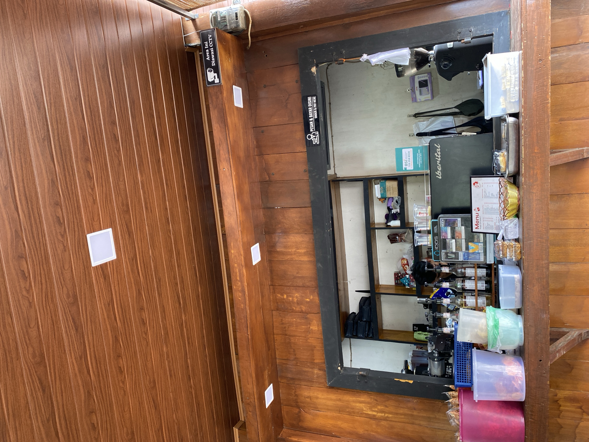 The image depicts the interior of a cafe in Bireuenmeunasahdayah, Indonesia, specifically a counter area. The counter is made of dark wood and is lined with various items, including plastic containers of different colors (pink, purple, light green), a metal tray with food, and a clear plastic container filled with biscuits. Behind the counter, there are shelves with bottles, a coffee grinder, a coffee machine labeled "iberital", and a menu board. The walls and ceiling are also made of wood, with recessed square lights visible in the ceiling. Signs are present on the wall and above the counter. One sign reads "Area Ini Diawasi CCTV" (This area is monitored by CCTV) with a camera icon. Another sign above the counter opening says "PESAN & BAYAR DISINI | ORDER & PAY HERE" with a pictogram of a person ordering. The overall impression is of a functional and somewhat rustic cafe setting. No people are clearly visible in the frame.
