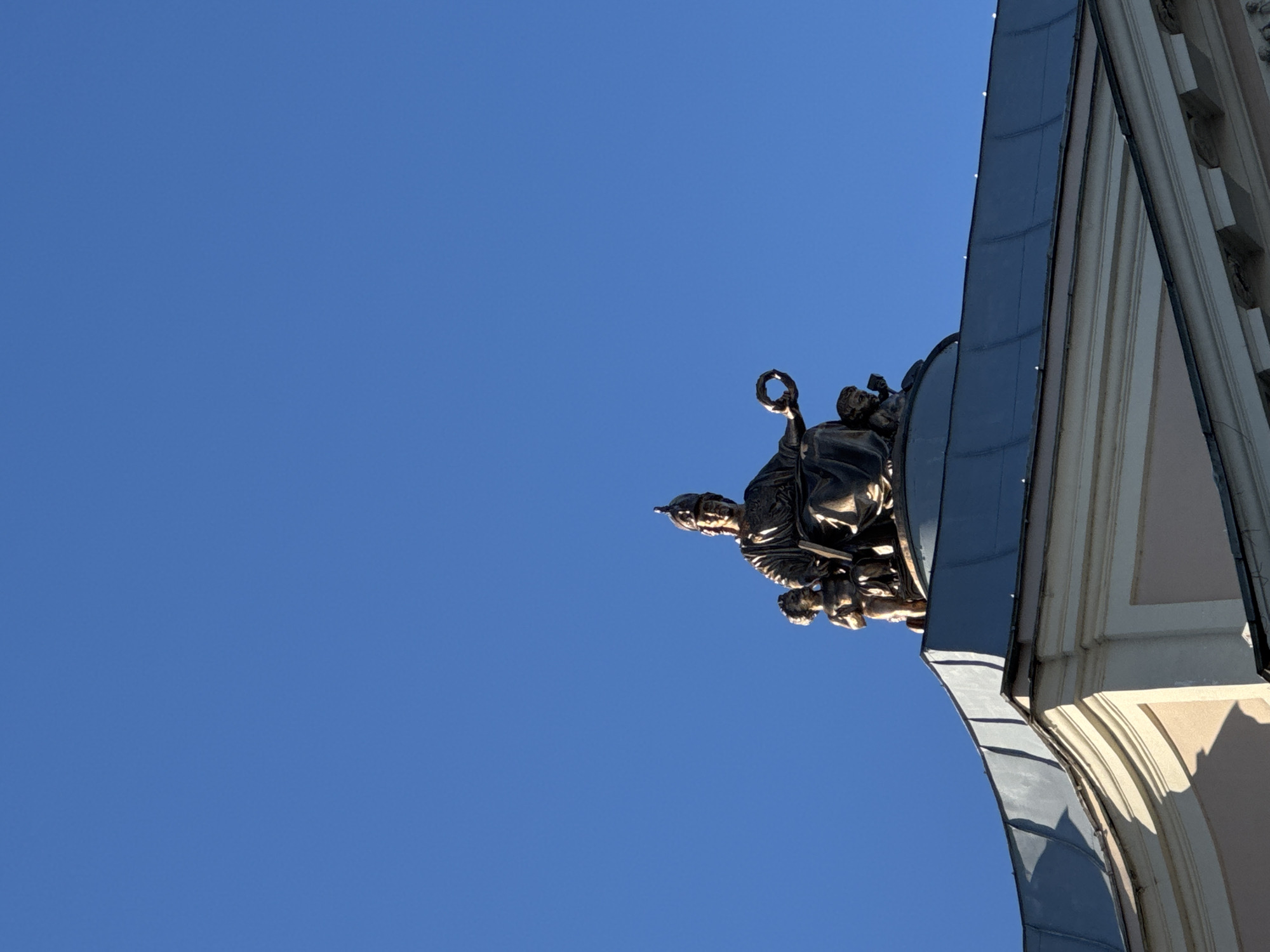 The image depicts a low-angle view of a section of a building against a clear blue sky. The building features an ornate architectural design with a statue or sculptural group positioned prominently on its roof or upper portion. The statue appears to depict a group of figures, possibly historical or allegorical. The building's facade is visible, showcasing details like moldings and decorative elements. The architecture is characterized by classical or baroque influences. Given the information about the location, the image is likely showcasing a detail of one of the historic buildings in Saint Petersburg, Russia, potentially a church, palace, or public building.